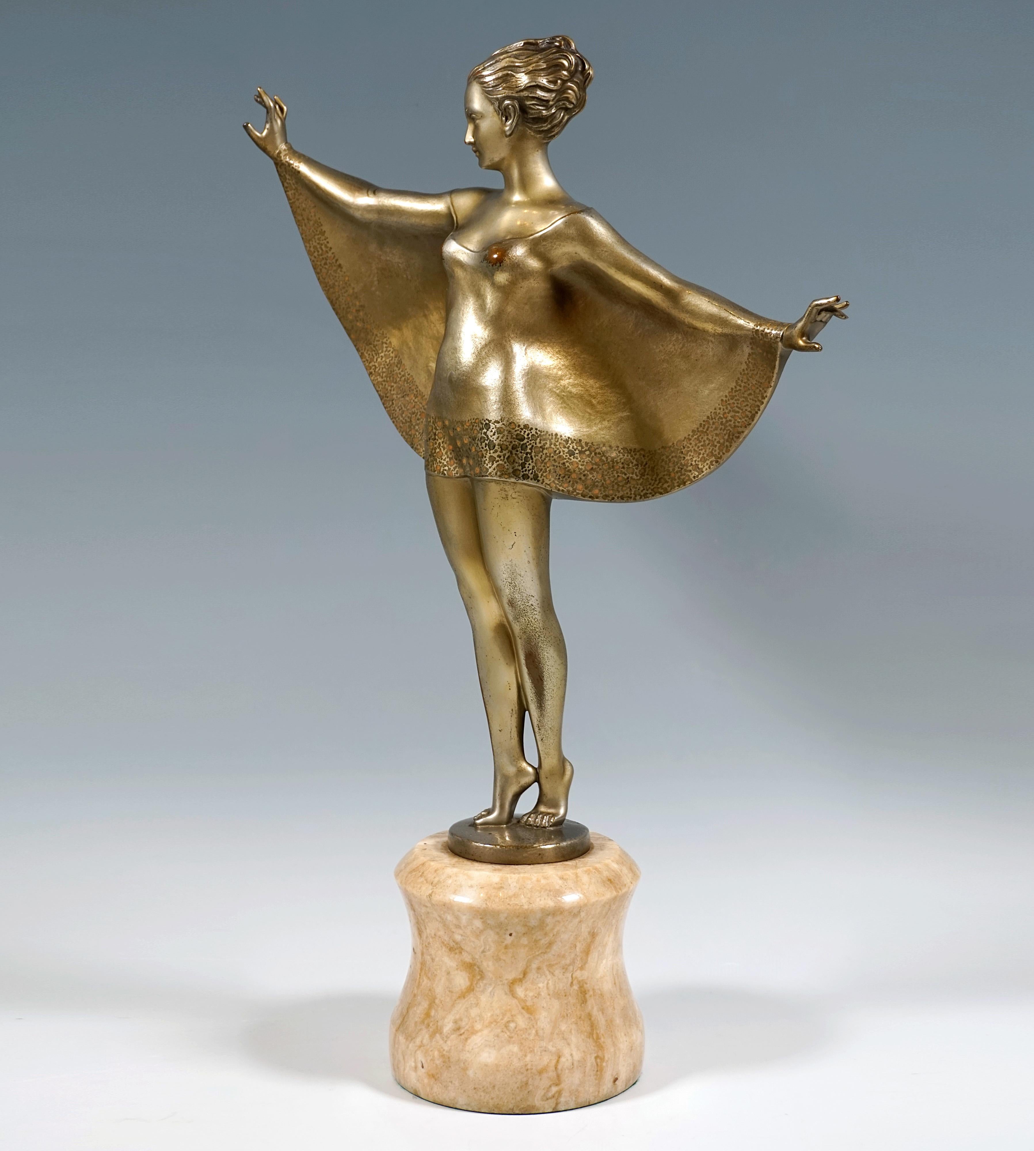 The petite dancer with short curly hair combed up high at the back balances on tiptoe, her upper body turned to the right and her arms stretched out horizontally from her body. She is wearing a short dress with batwing sleeves that extends in a