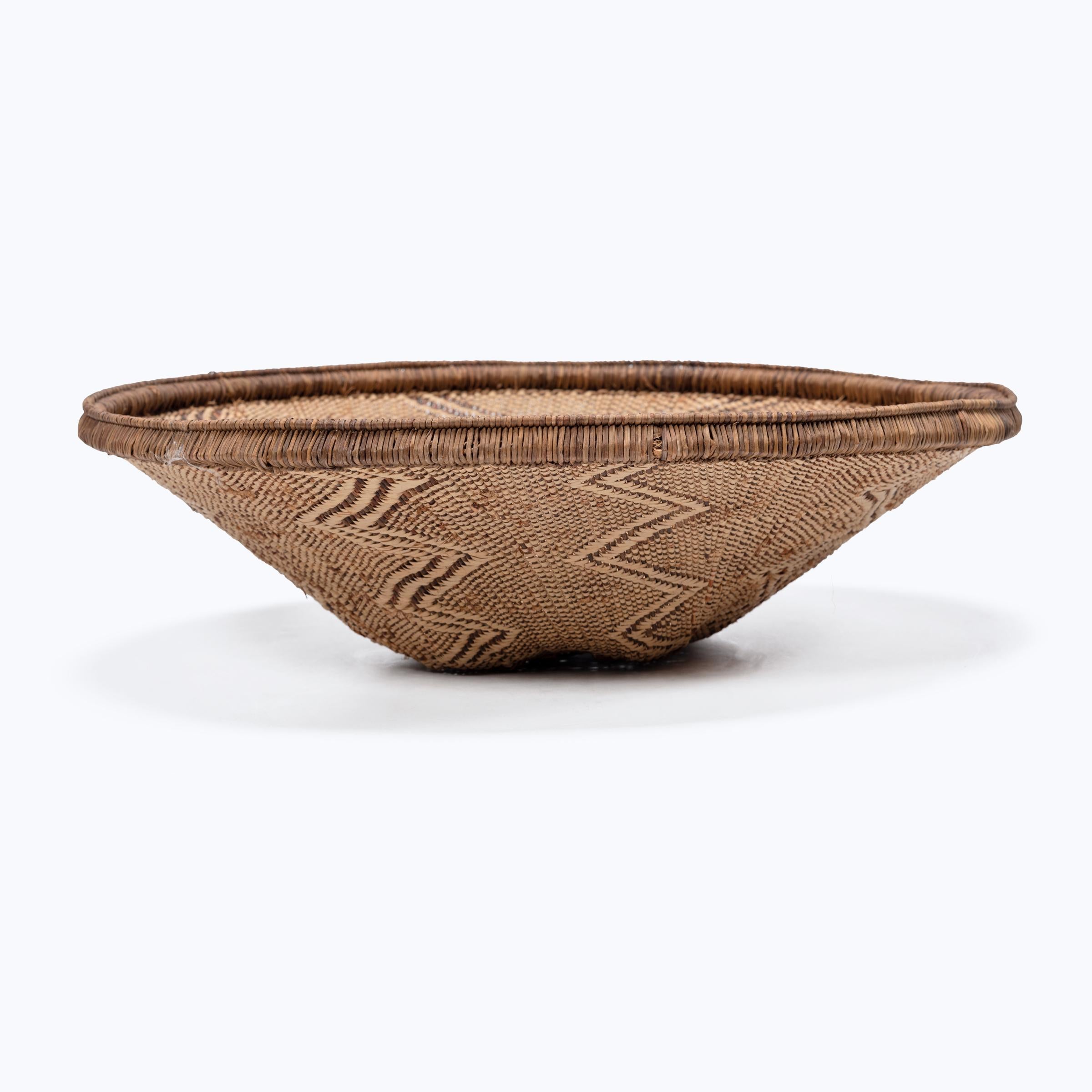Tightly woven of reeds and willow, this round tray basket was crafted by artisans of the baTonga people of Zambia and Zimbabwe. Contrasting light and dark materials create zig-zag patterns that run down the basket's tapered form, ending in a flat