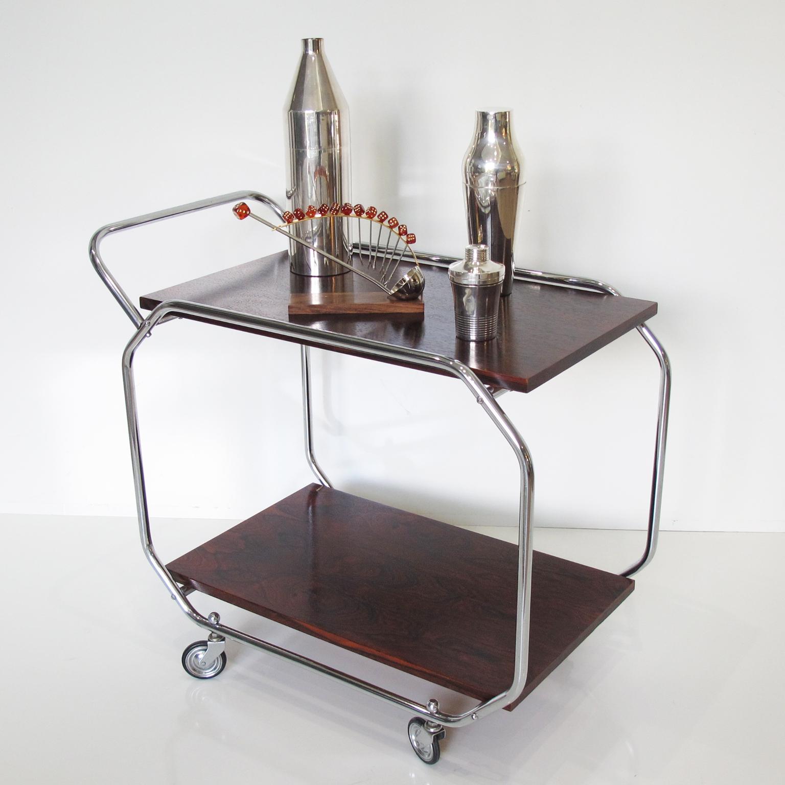 This striking, streamlined Art Deco rolling bar serving cart was designed with the influence of Bauhaus Modernism. This bar accessory features a chromed metal round tubing frame with wood upper and lower shelves. The piece boasts a sturdy chrome