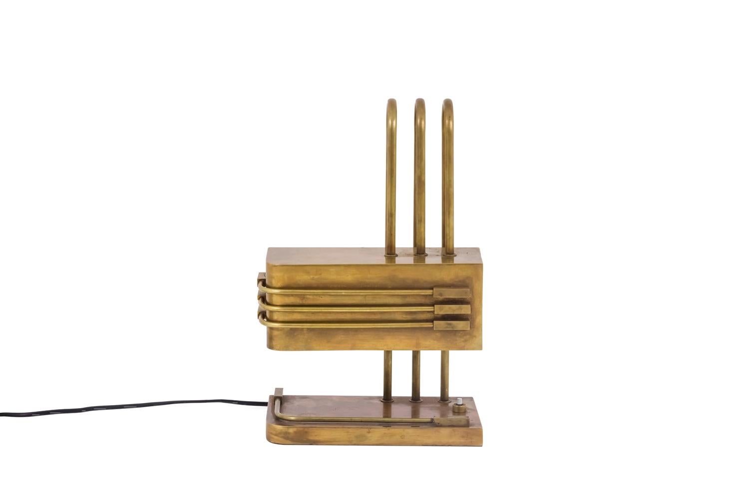 Bauhaus lamp in patinated gilt brass standing on a rectangular base with a curved angle, decorated with an applied form following the curved angle.
Shaft composed of three gilt bronze sticks slightly spaced forming an arch.
Rectangular lampshade