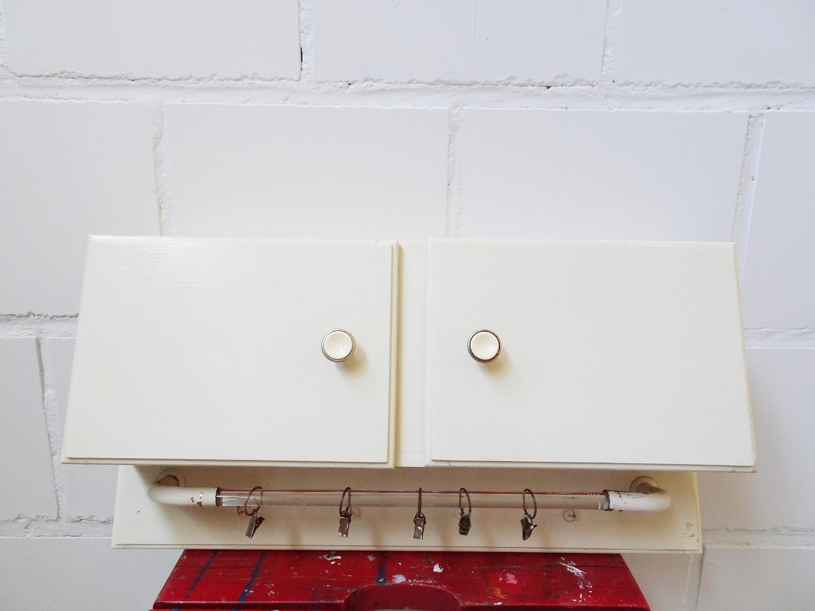 This small wall cabinet is a kitchen cabinet from the early 1930s. The cream white softwood wall cabinet features a spacious interior with two bakelite doorhandles, as well as a large glass hook rack with five clips and four additional hooks. A