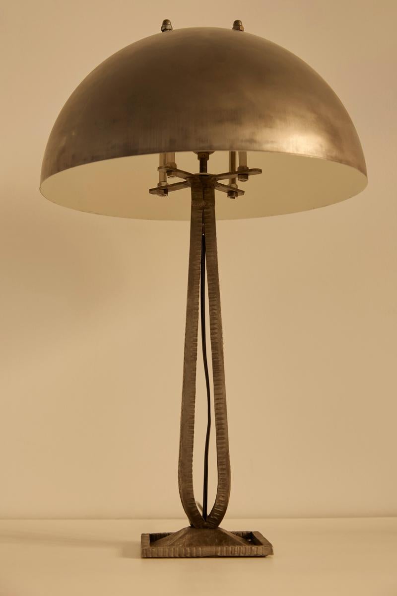 Esta lámpara de sobremesa presenta una pantalla metálica en forma de cúpula, una característica base martillada y líneas elegantes y geométricas que evocan el diseño inspirado en la Bauhaus. La combinación de materiales industriales y detalles