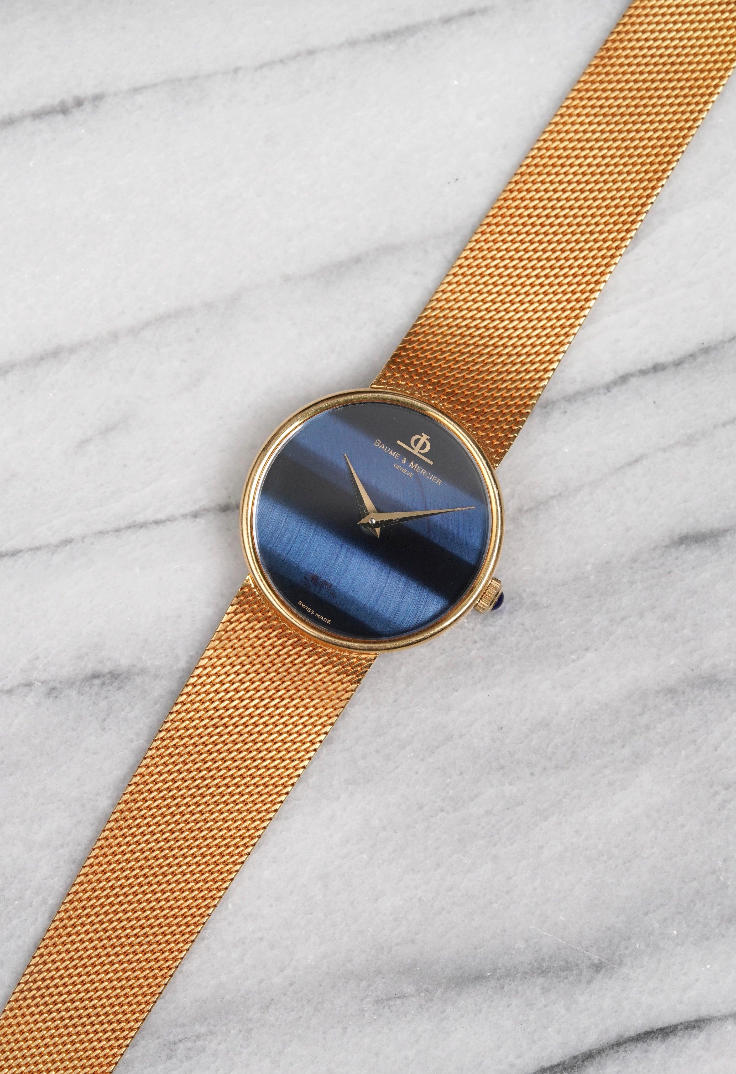Ecco un Baume & Mercier in oro giallo 18 carati con quadrante in pietra blu occhio di falco a carica manuale degli anni Settanta. Le misure includono una cassa di 33 mm e un bracciale integrato che si adatta a un polso di 7 pollici. Solo tempo. Le