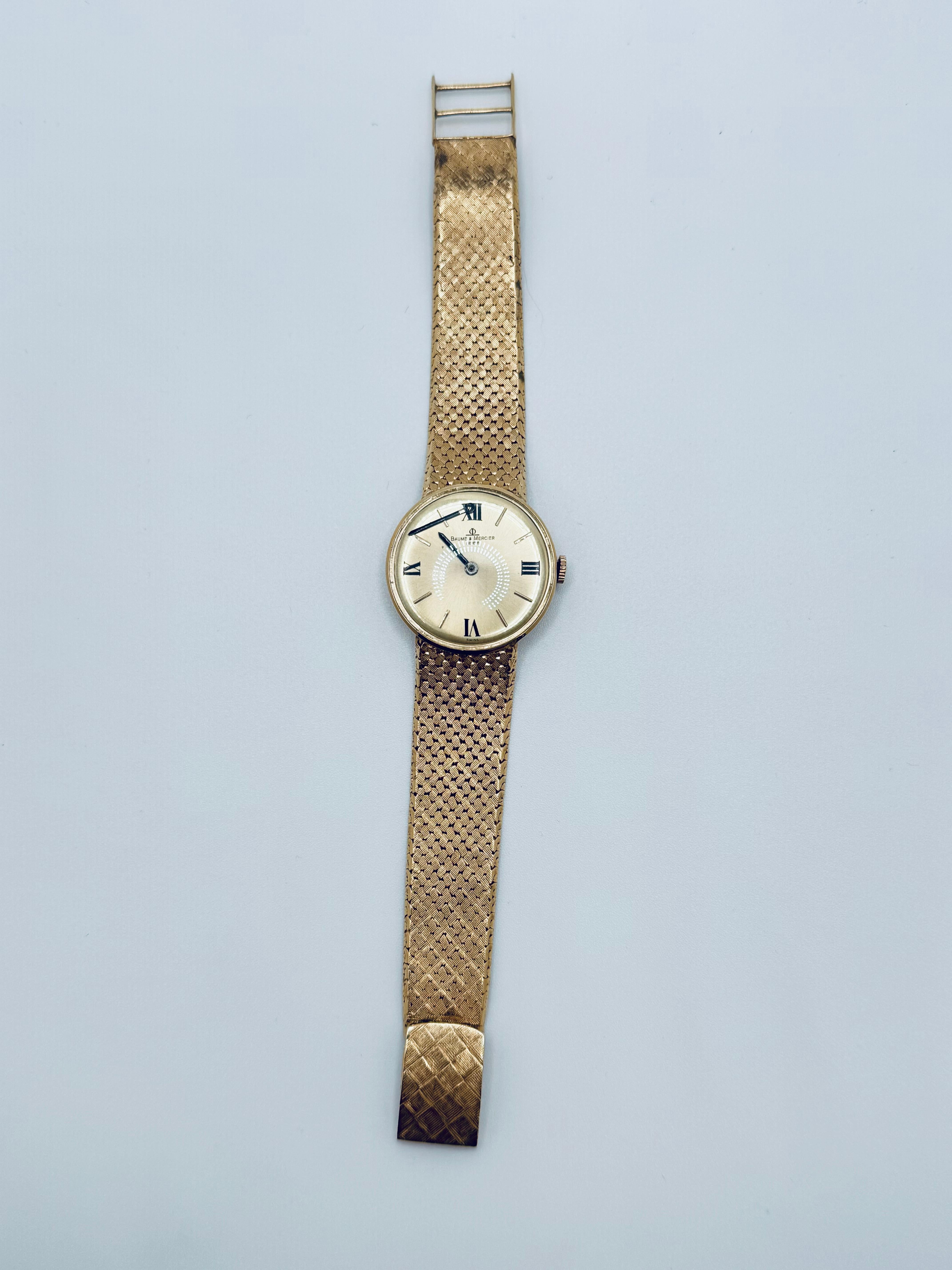 This Baume & Mercier Genève men’s watch showcases a refined design in yellow gold, featuring a wide mesh-finish band that offers both comfort and elegance. The dial and bezel are crafted in matching yellow gold, creating a seamless and sophisticated