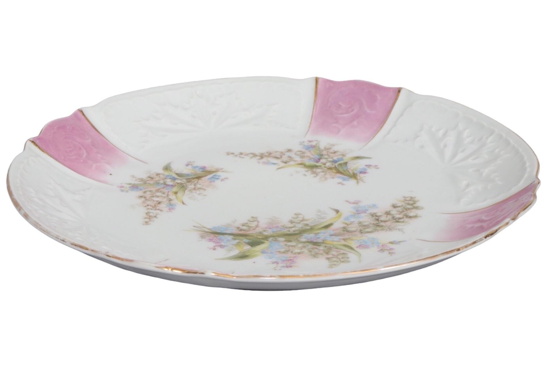 A Bavarian style ceramic serving plate. Hand painted in the center with wild violets and ivory bells. The outer band is embossed with holly leaves and roses, with bands of iridescent pink and trimmed with a gilt line along the serpentine edge.