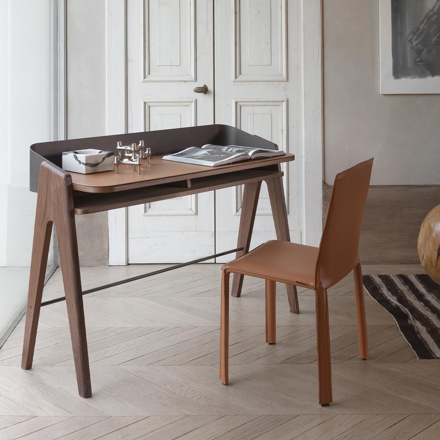 The Bavero desk combines an elegant, curved metal frame with a smooth cognac leather pad, creating a seamless design without visible screws. This desk features a wooden structure with slender walnut legs and includes a practical shelf beneath the