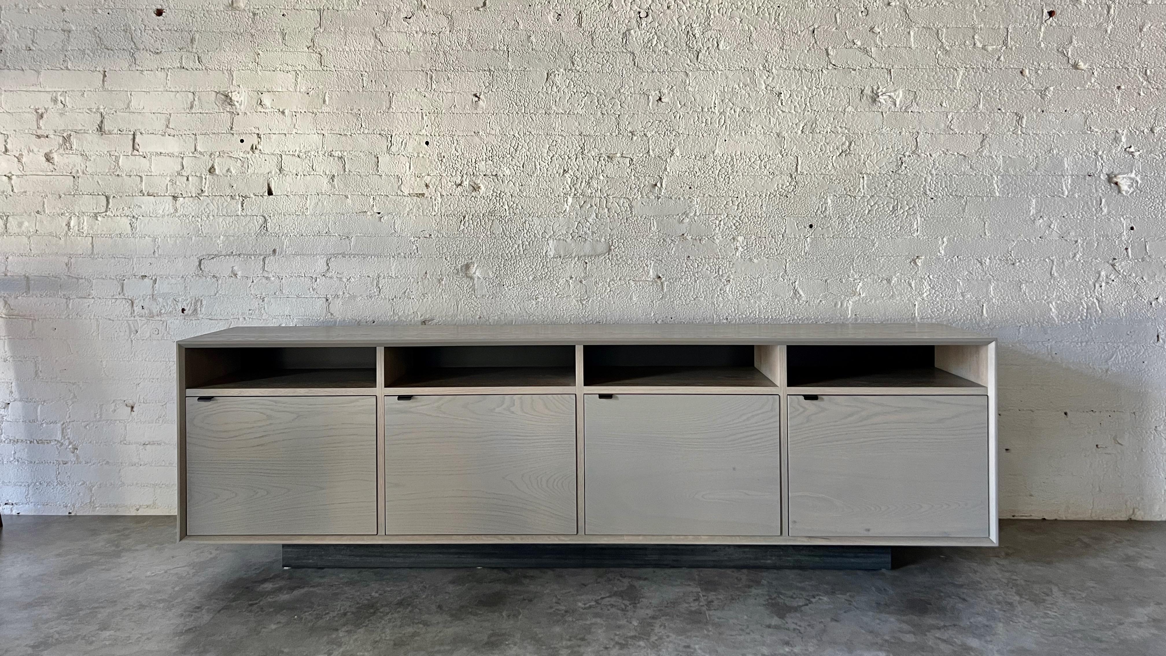 The Baxter Credenza has a low and lean profile, showcasing four cabinet doors and open upper storage. The credenza uses solid white oak constructed with, an uncommonly practiced, entirely solid wood carcass and cabinet doors. Black steel door pulls,