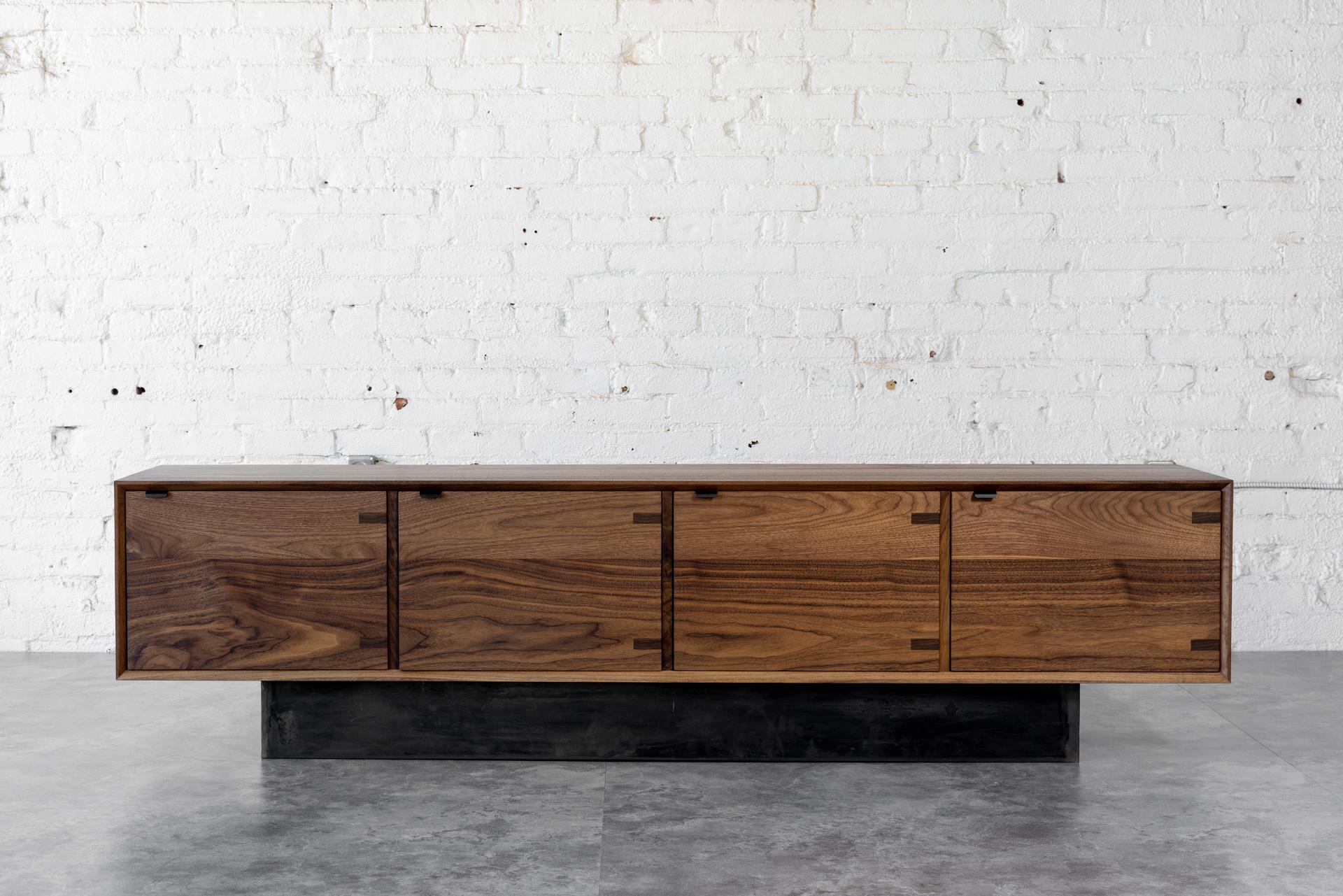 The Baxter walnut low Credenza has a sleek profile, showcasing four cabinet doors. The credenza uses all solid black walnut constructed with, an uncommonly practiced, entirely solid wood carcass, interior shelves, and credenza doors. Black steel