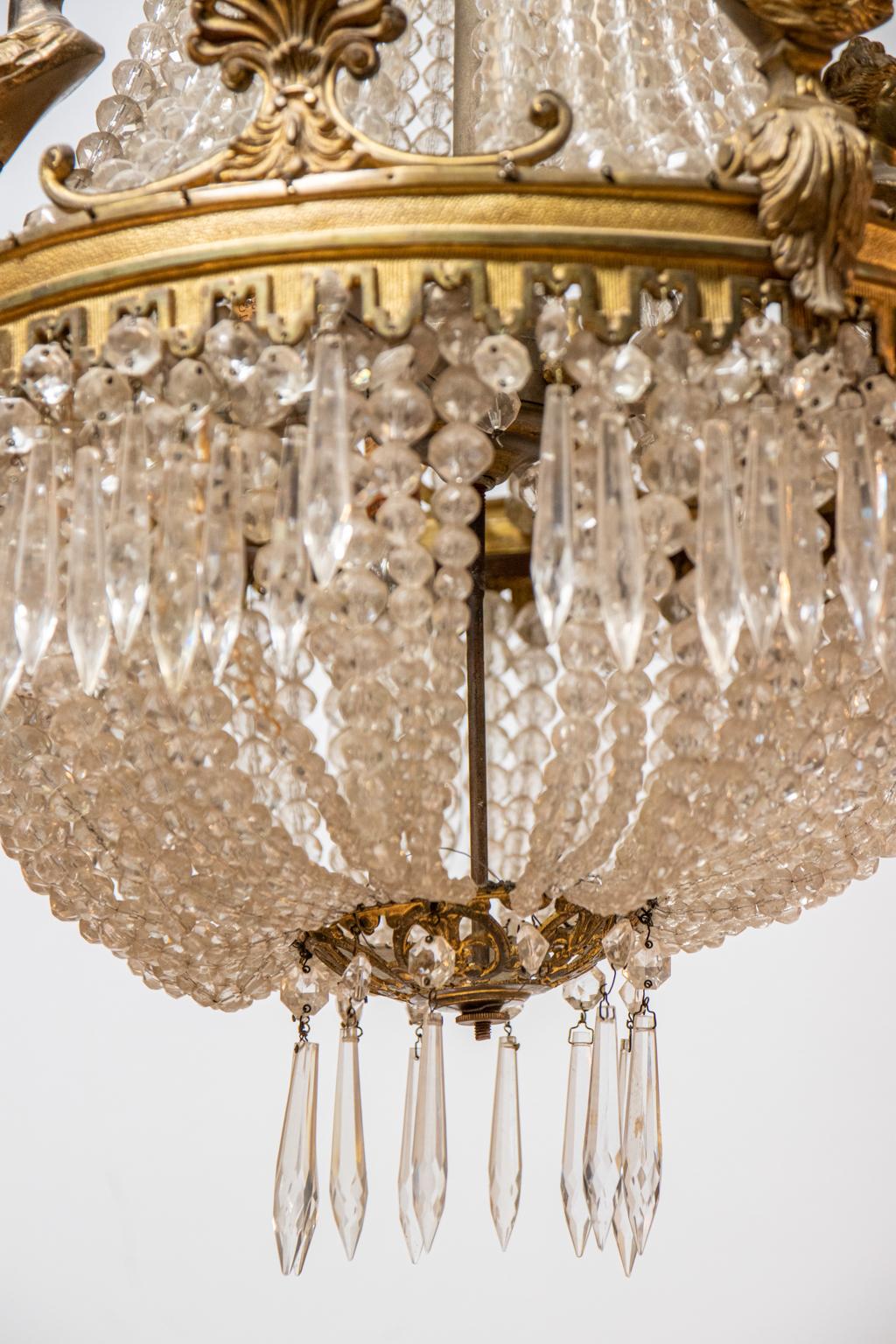 Beaded crystal chandelier with metal palmettes and stylized female half figure motifs. The chandelier is also constructed with scrolled foliage accents, pointed drop shaped crystal prisms, and beaded crystal swags. The top canopy is further detailed