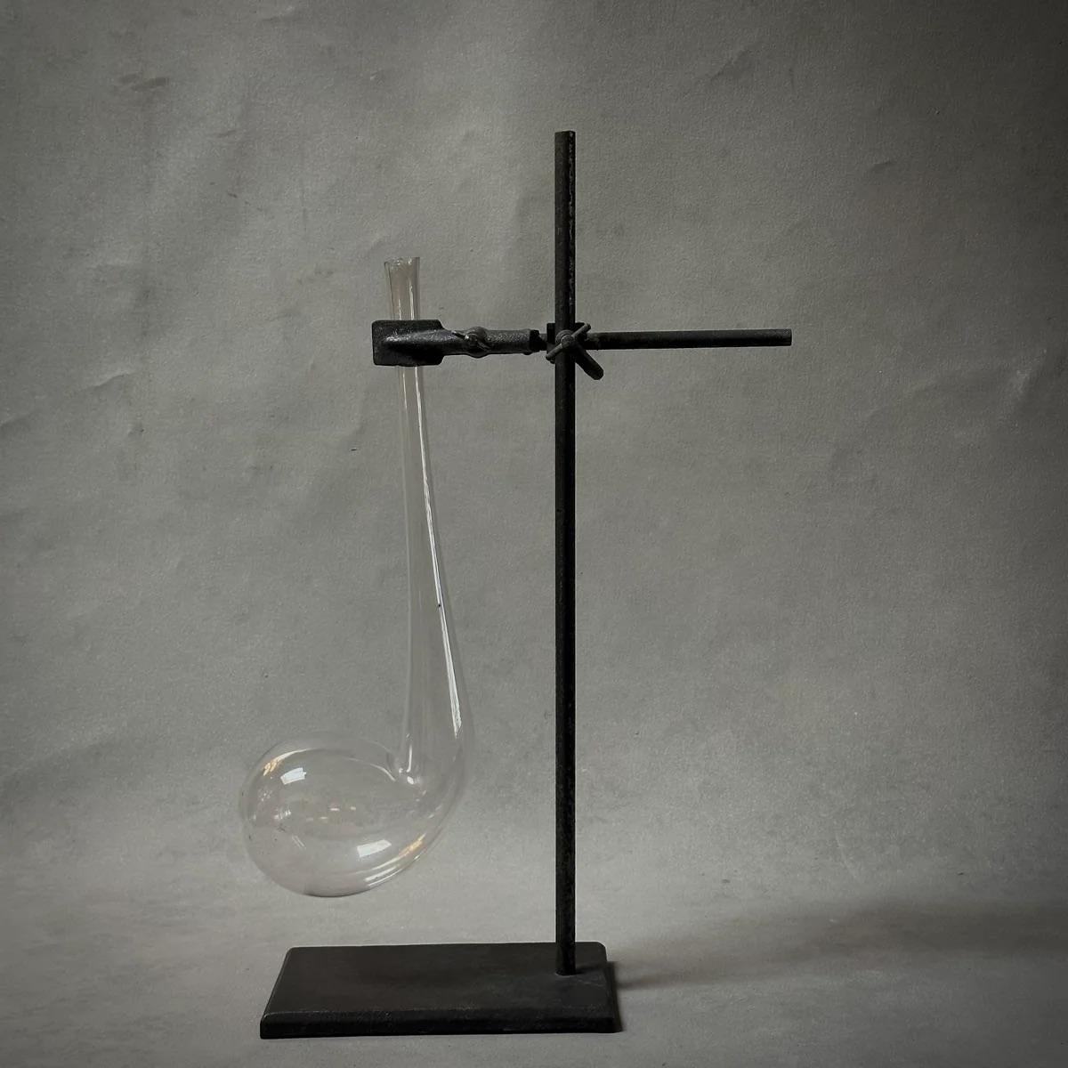 This early 20th-century Belgian laboratory beaker, complete with its original metal stand, captures a moment in scientific history. The glass, shaped like a pipe, is delicately thin, creating a striking contrast with its robust, utilitarian metal