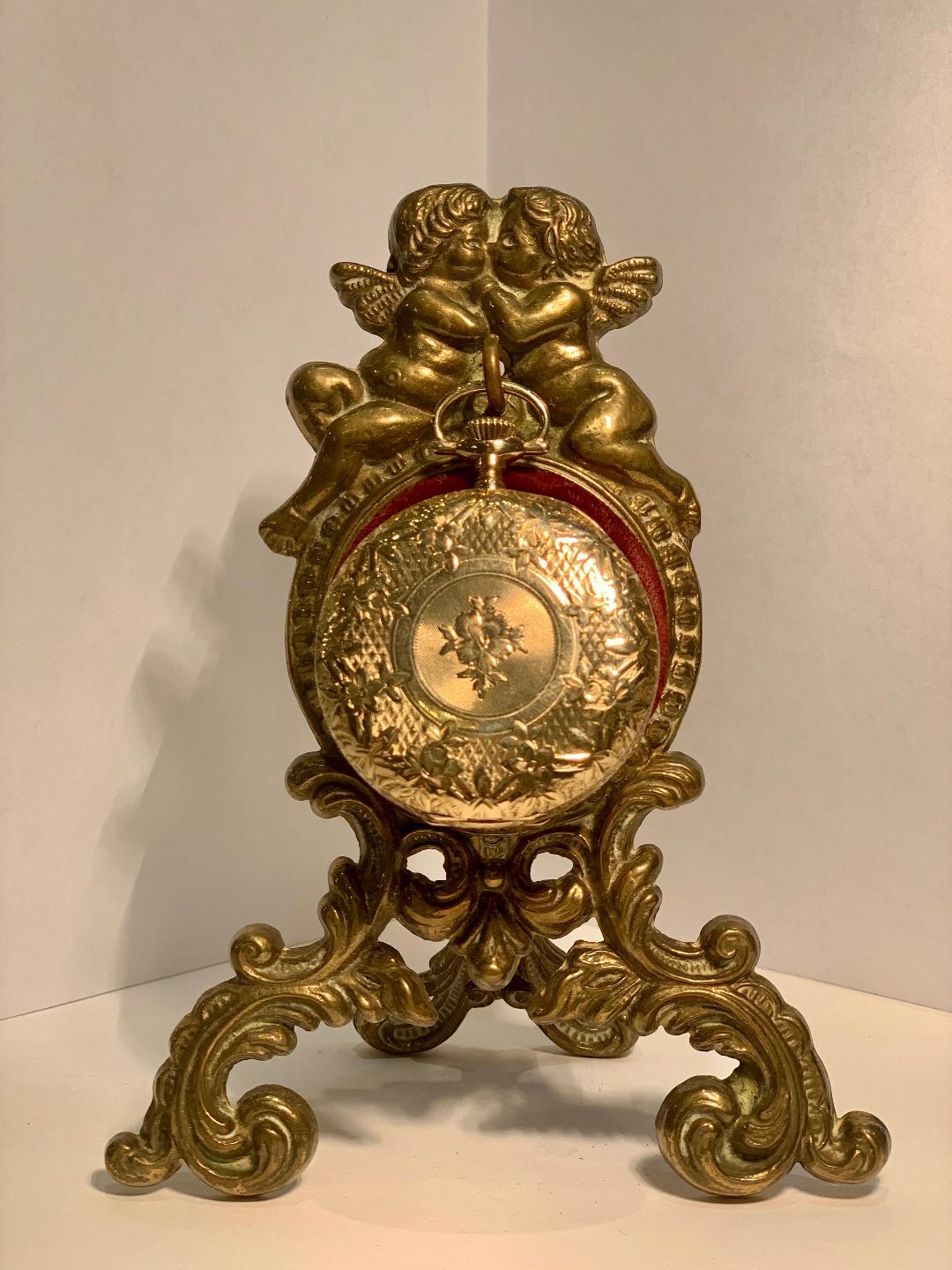 Beautifully handcrafted antique display stand or porte montre from which to hang a pocket watch features an ornate, baroque scrolling motif antique gold finish metal frame with a pair of loving cherubs on top and a burgundy velvet circular cushion