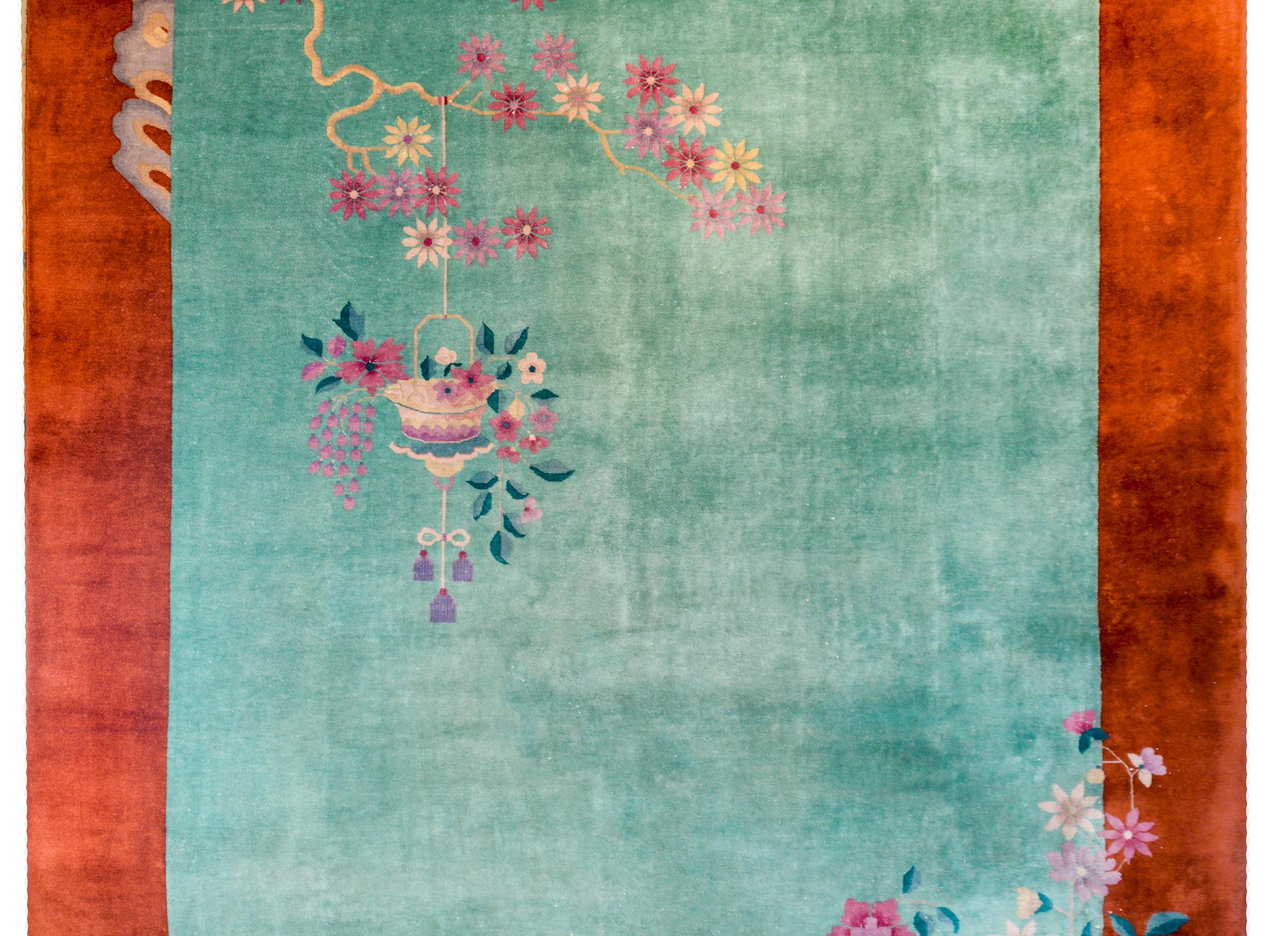 A beautiful early 20th century Chinese Art Deco rug with a gorgeous sea-foam green field surrounded by a wide rust orange border. A potted peony lives in one corner while a gnarly twisted flowering prunus branch with a hanging basket of flowers