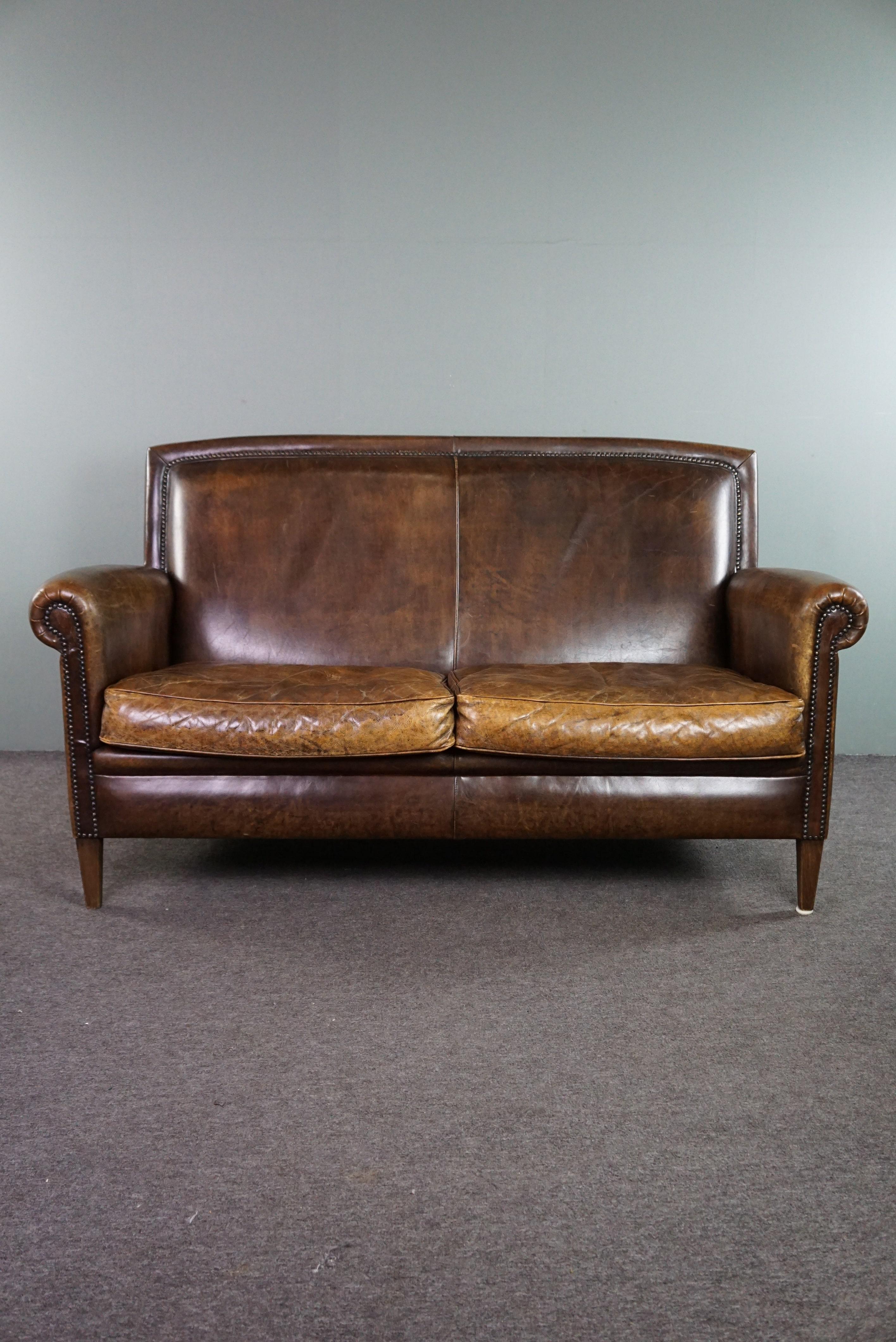 Offered: this beautiful and well-maintained dark cognac-colored cowhide 2-seater sofa in classic English style, finished with decorative studs.

This lovely sofa has a solid and dignified appearance, and it feels just as good as it looks. The