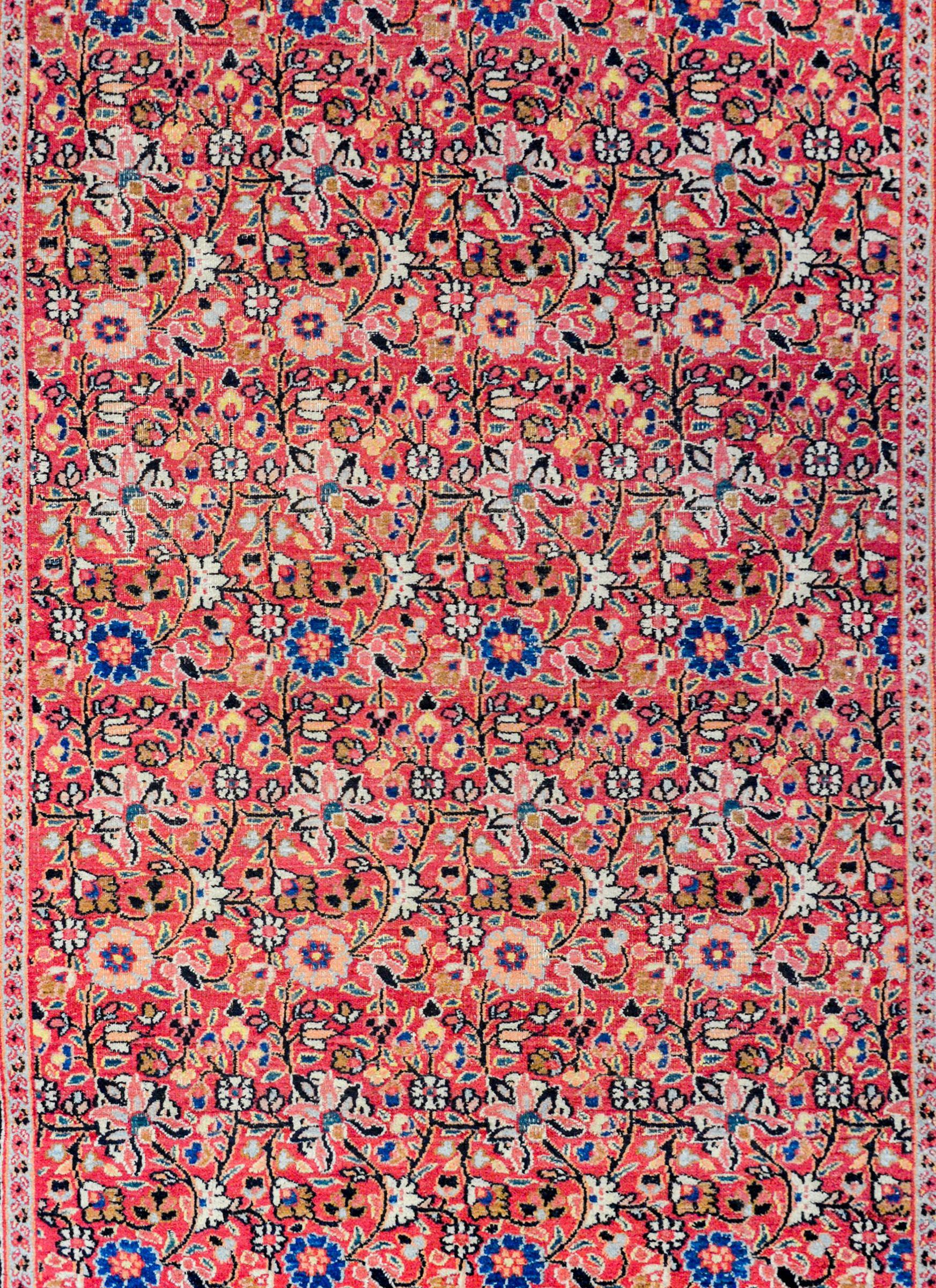 A beautiful early 20th century Persian Khorasan rug with an all-over repeated pattern containing myriad varieties of flowers woven in light and dark indigo, cranberry orange, green, pink, and beige vegetable dyed wool on a cranberry field surrounded