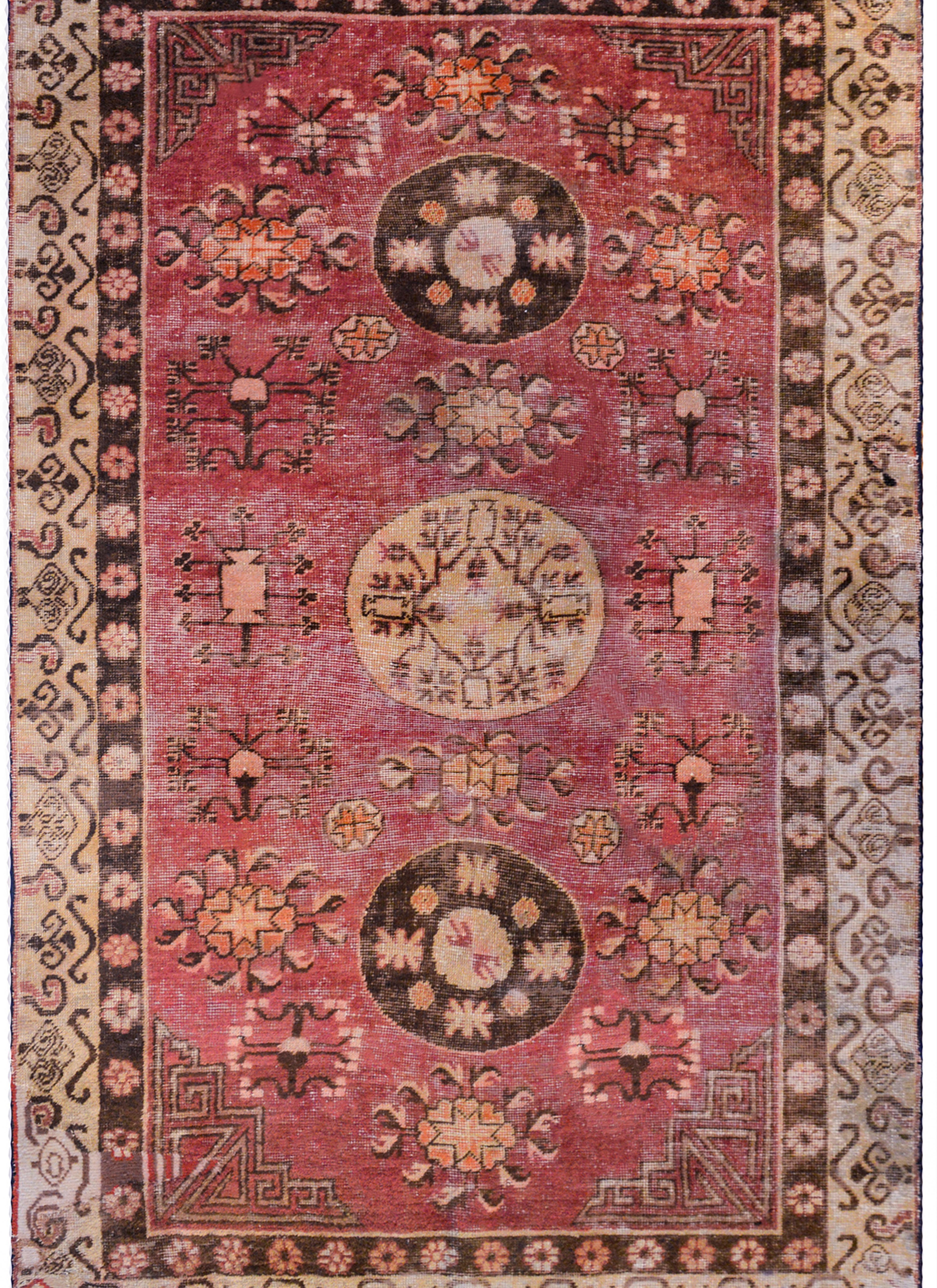 A beautiful early 20th century Central Asian Khotan rug with three round medallions each with a stylized floral pattern, amidst a field of trees-of-life on a dark cranberry field. The border is composed with two patterned borders, one with a petite