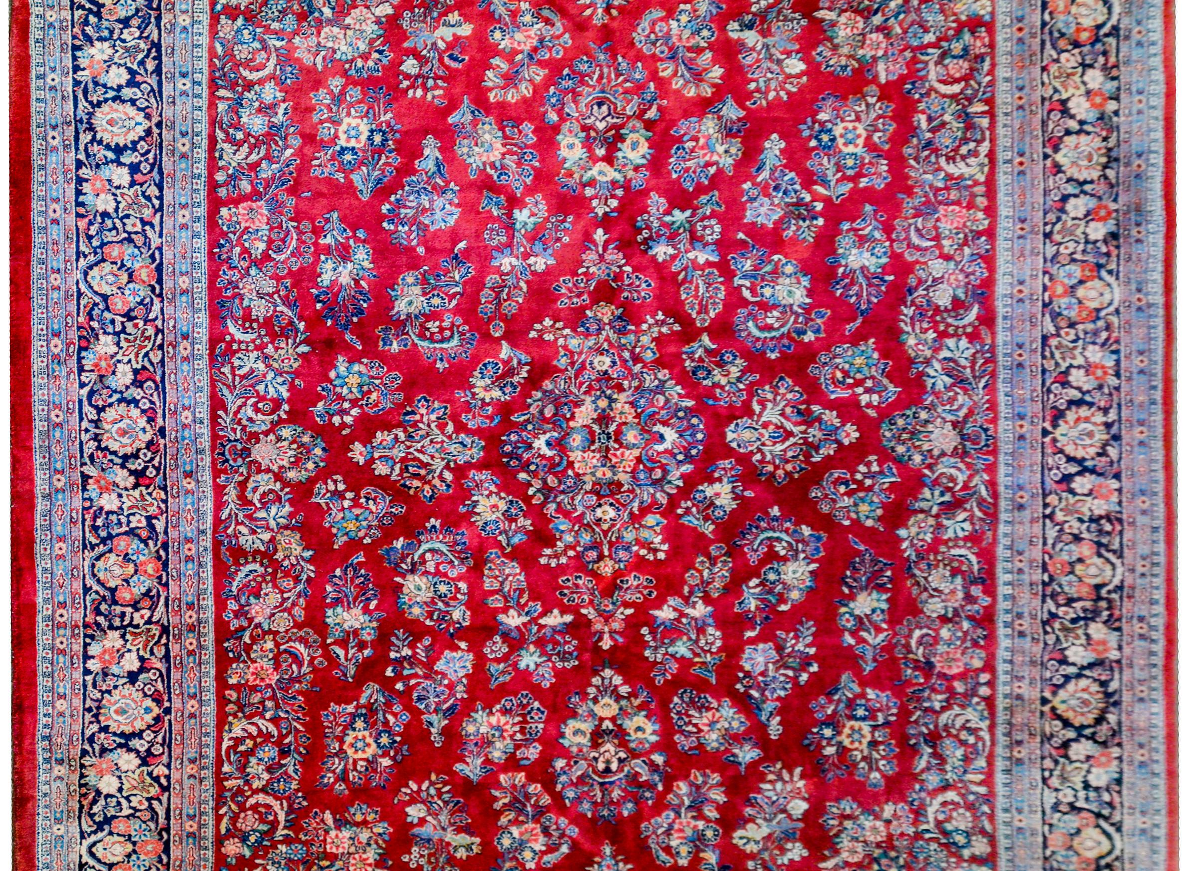 A beautiful early 20th century Persian Sarouk rug with an amazing mirrored myriad floral and leaf pattern woven in light and dark indigo, gold, and pink colored wool on a deep crimson background. The border is complex with a wide, densely woven,