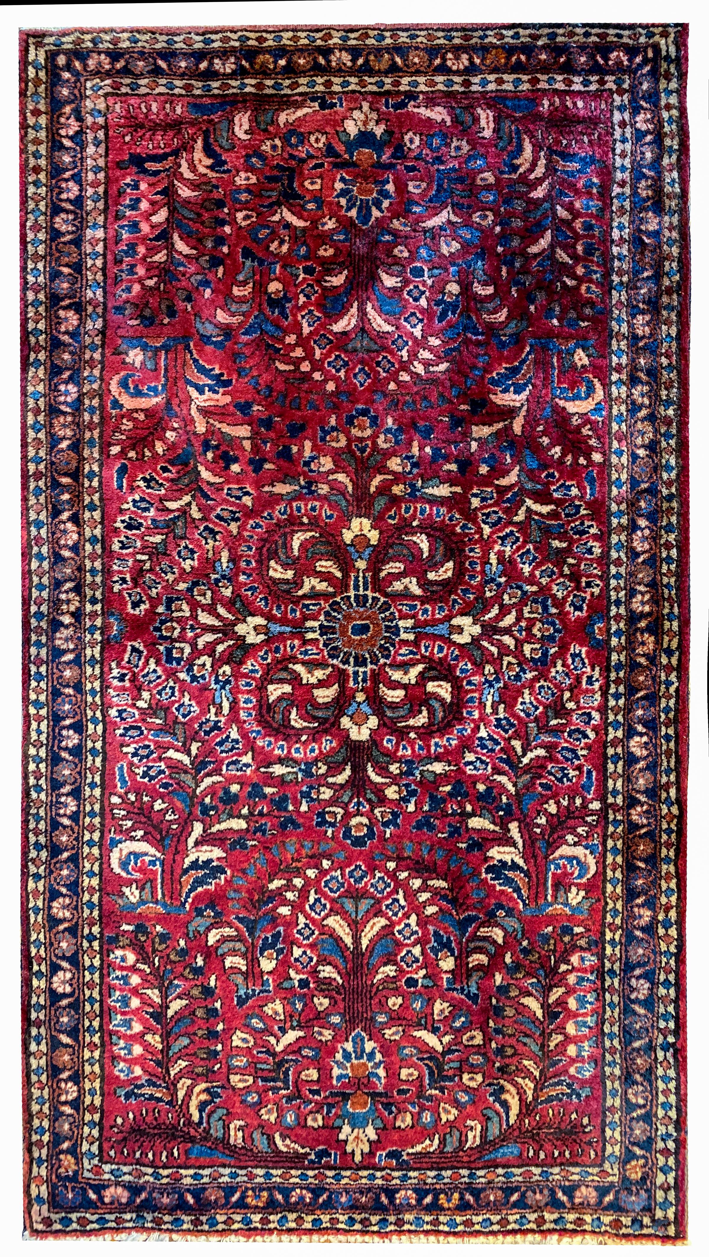 A beautiful early 20th century Persian Sarouk rug with a traditional mirrored tree-of-life pattern including myriad flowers and scrolling vines woven in light and dark indigo, cream, green, and brown, on a bold dark cranberry background. The border