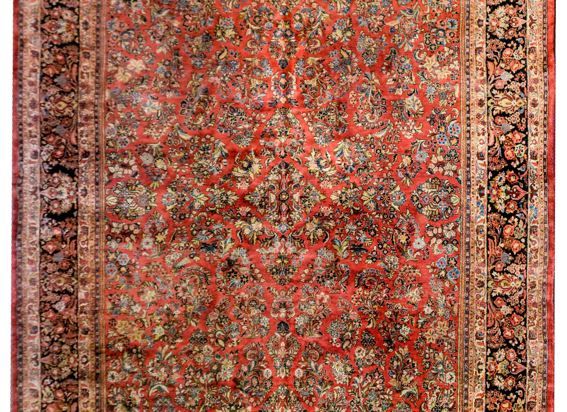 A beautiful early 20th century Persian Sarouk rug with a traditional all-over mirrored floral and vine tree-of-life pattern woven in light and dark indigo, gold, and cream colored wool, on a rich coral background. The border is complex with a