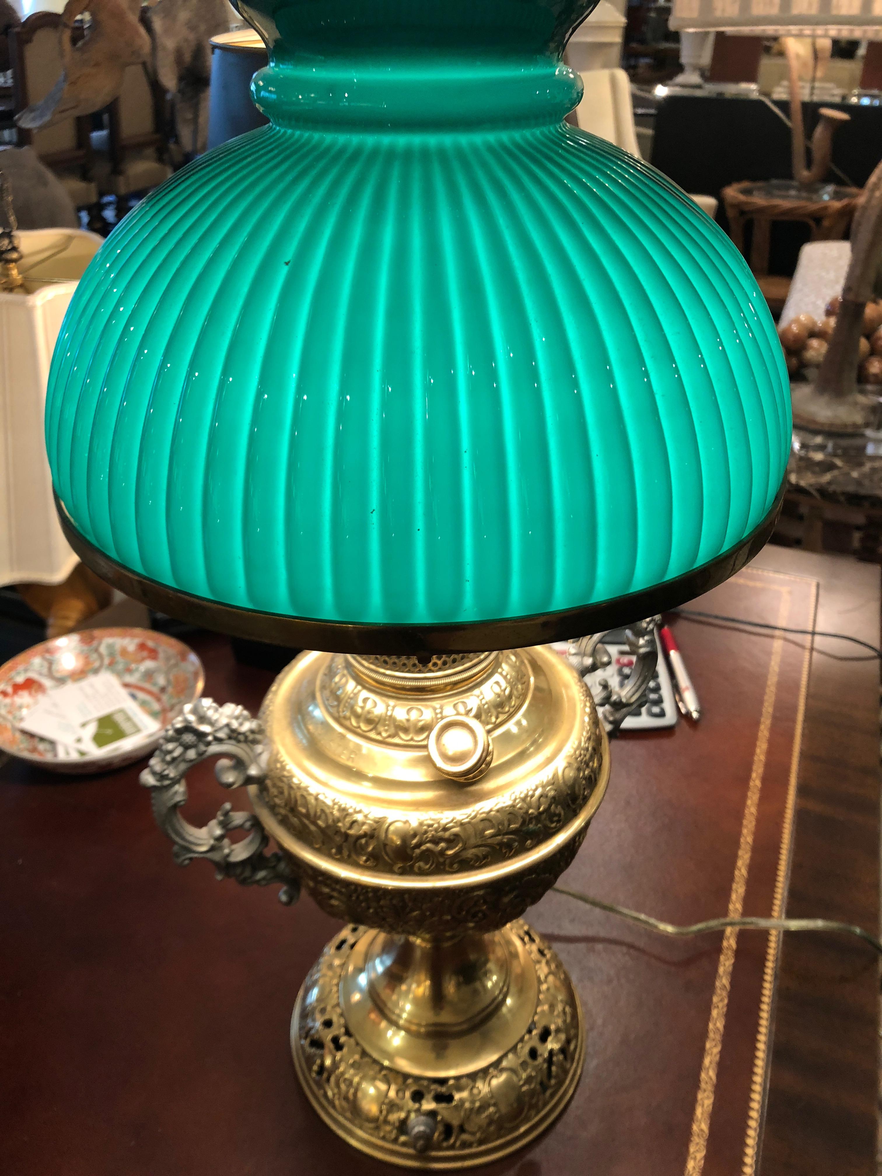 Ornately embossed brass electrified oil lamp with silver plate handles and New Rochester written on the side. There is a glass interior hurricane and gorgeous ribbed green glass shade, beautiful when unlit as well as when it's glowing bright.