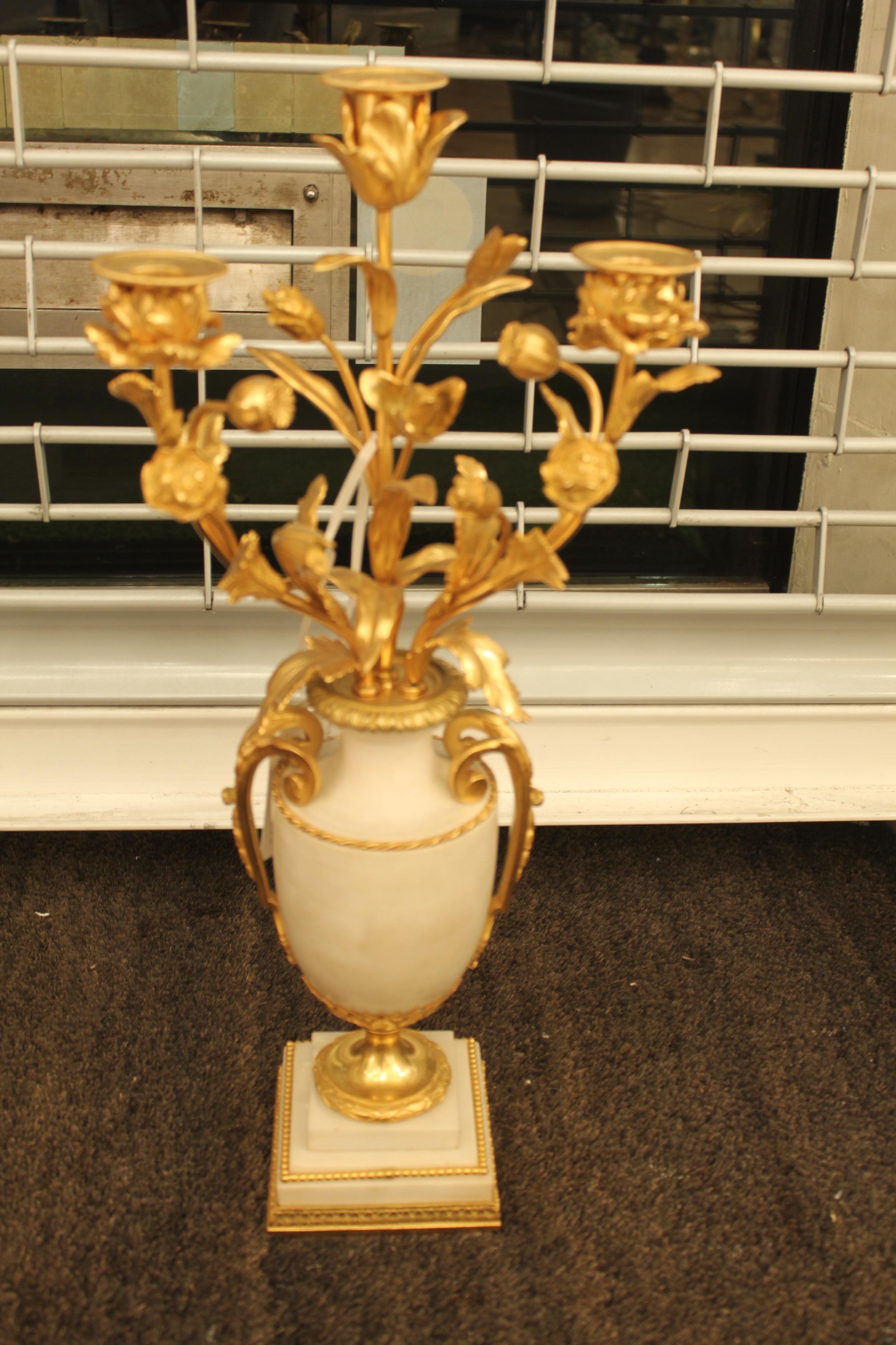 A beautiful pair of gilt bronze and marble candelabras on square decorative marble bases. Each holds three candles on floral designed stems surrounded by decorative foliage. The marble centre features either side two gilt bronze carrying handles