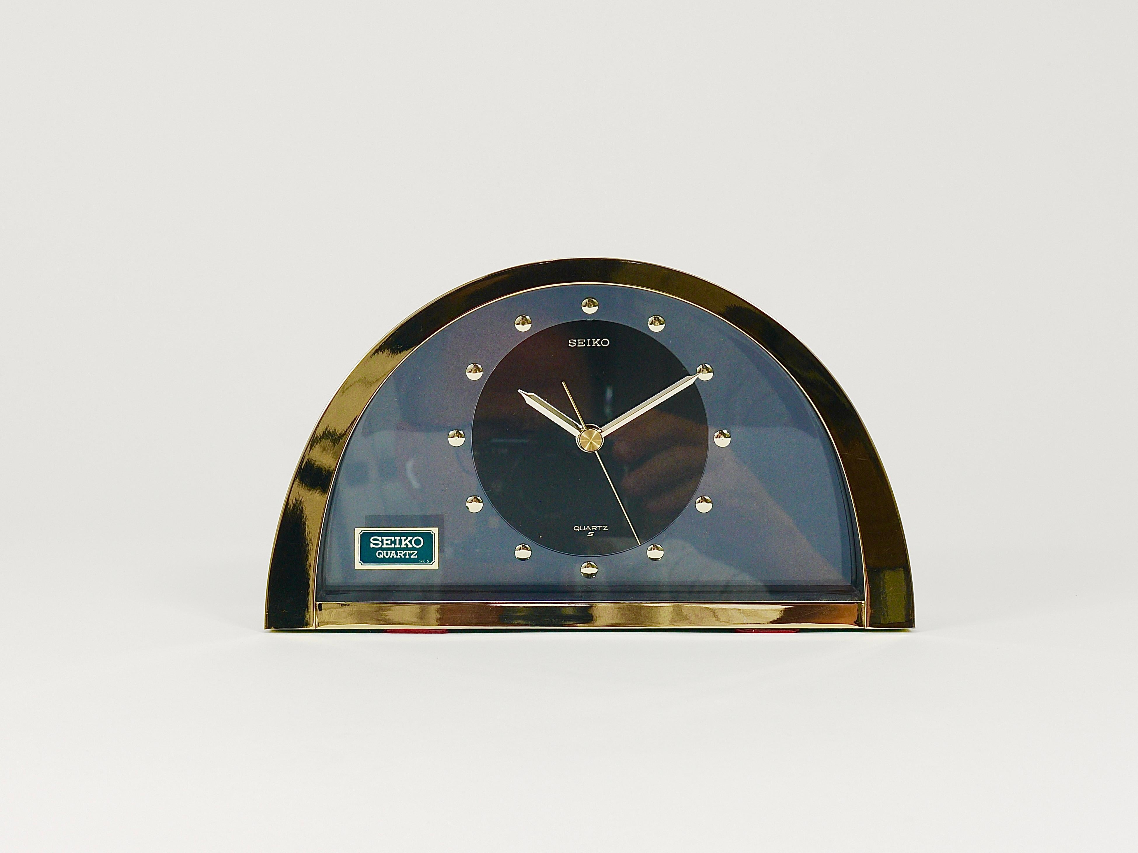 A decorative Hollywood Regency desk/table/mantel clock from the 1980s. It features a brushed brass housing with a mirror-polished front, elegant golden handles and indices, and a smoked-grey transparent 