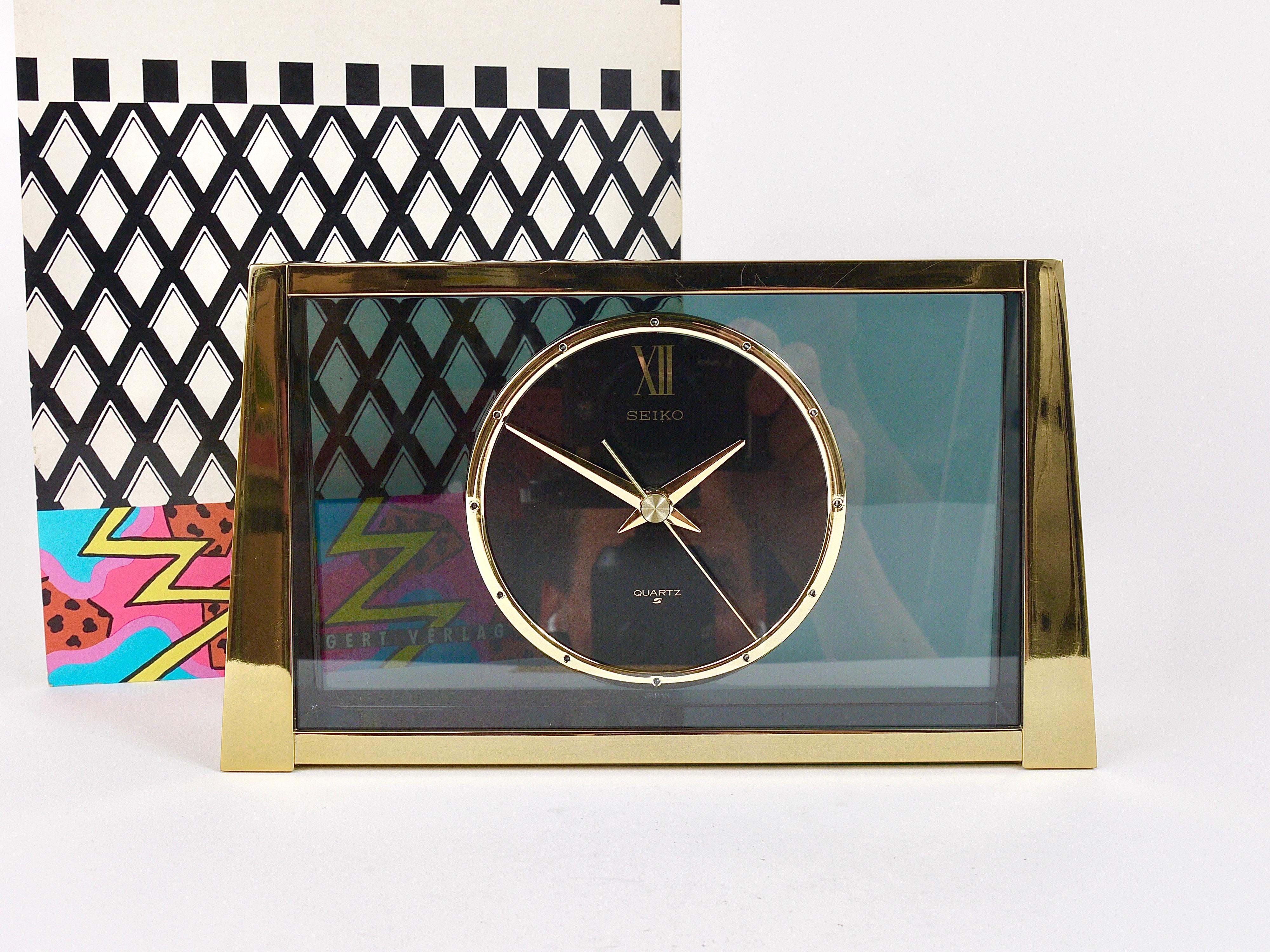 A decorative Hollywood Regency desk / table / mantel clock from the 1980s. It has a brushed brass housing with mirror polished front, nice golden handles and indices and a smoked-grey transparent 