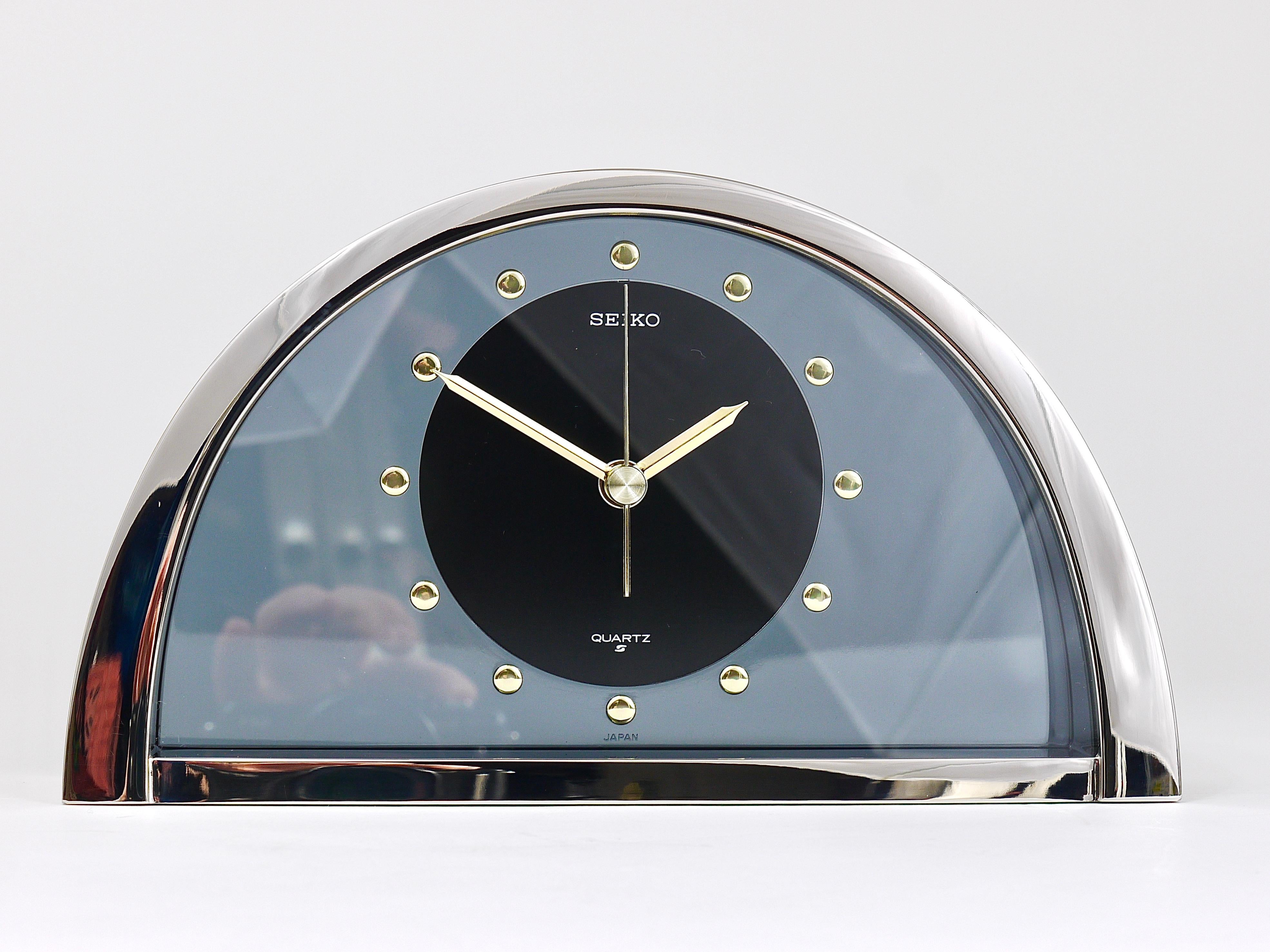 A decorative Hollywood Regency desk / table / mantel clock from the 1980s. It has a brushed chrome-plated housing with mirror polished front, nice golden handles and indices and a smoked-grey transparent 