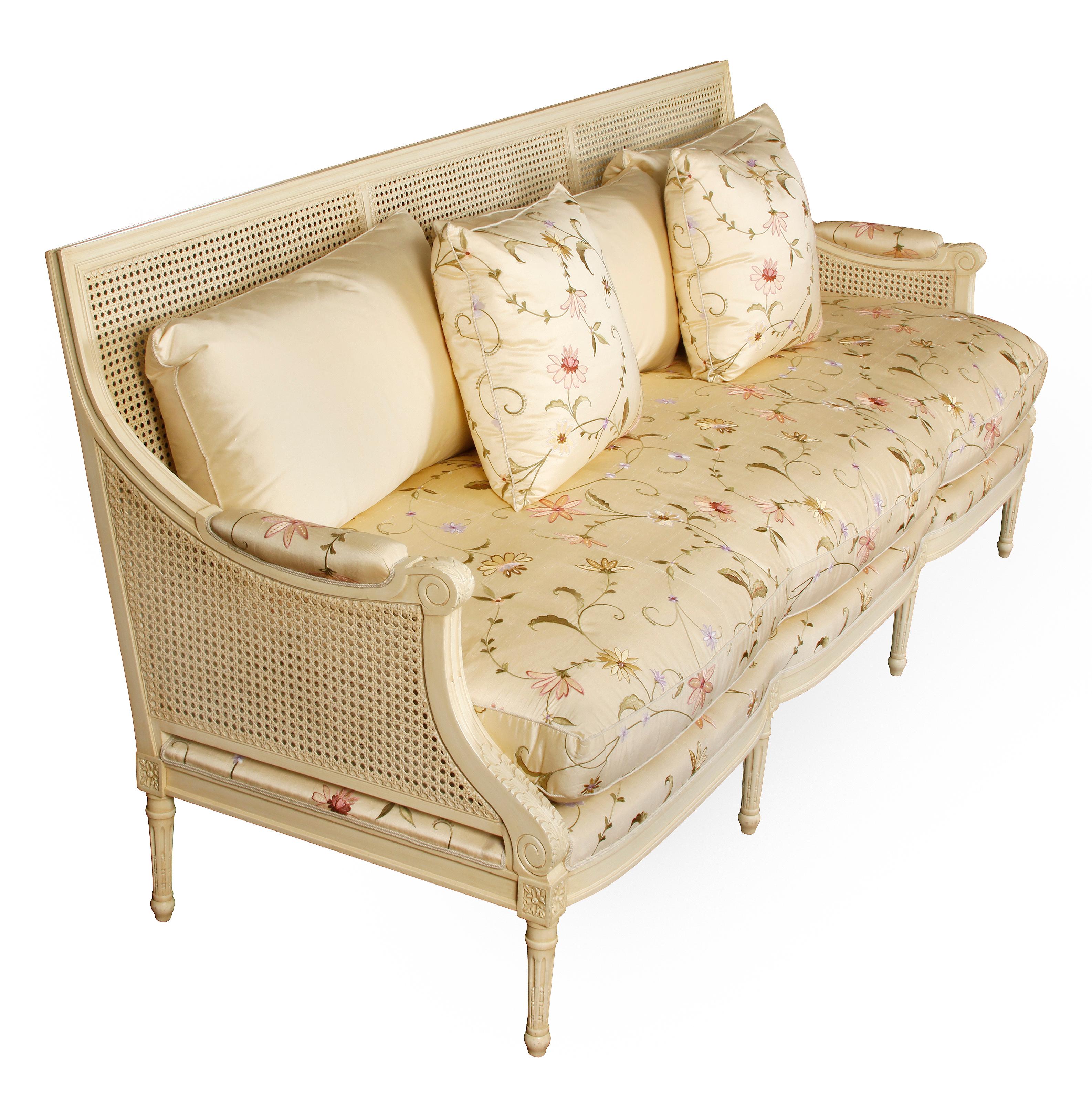 Beautiful painted ivory settee with cane back and sides, silk floral fabric seat cushion and pillows and ivory silk back cushions. Carved in Louis XVI style with Vitruvian scroll and floral details on arms and straight legs