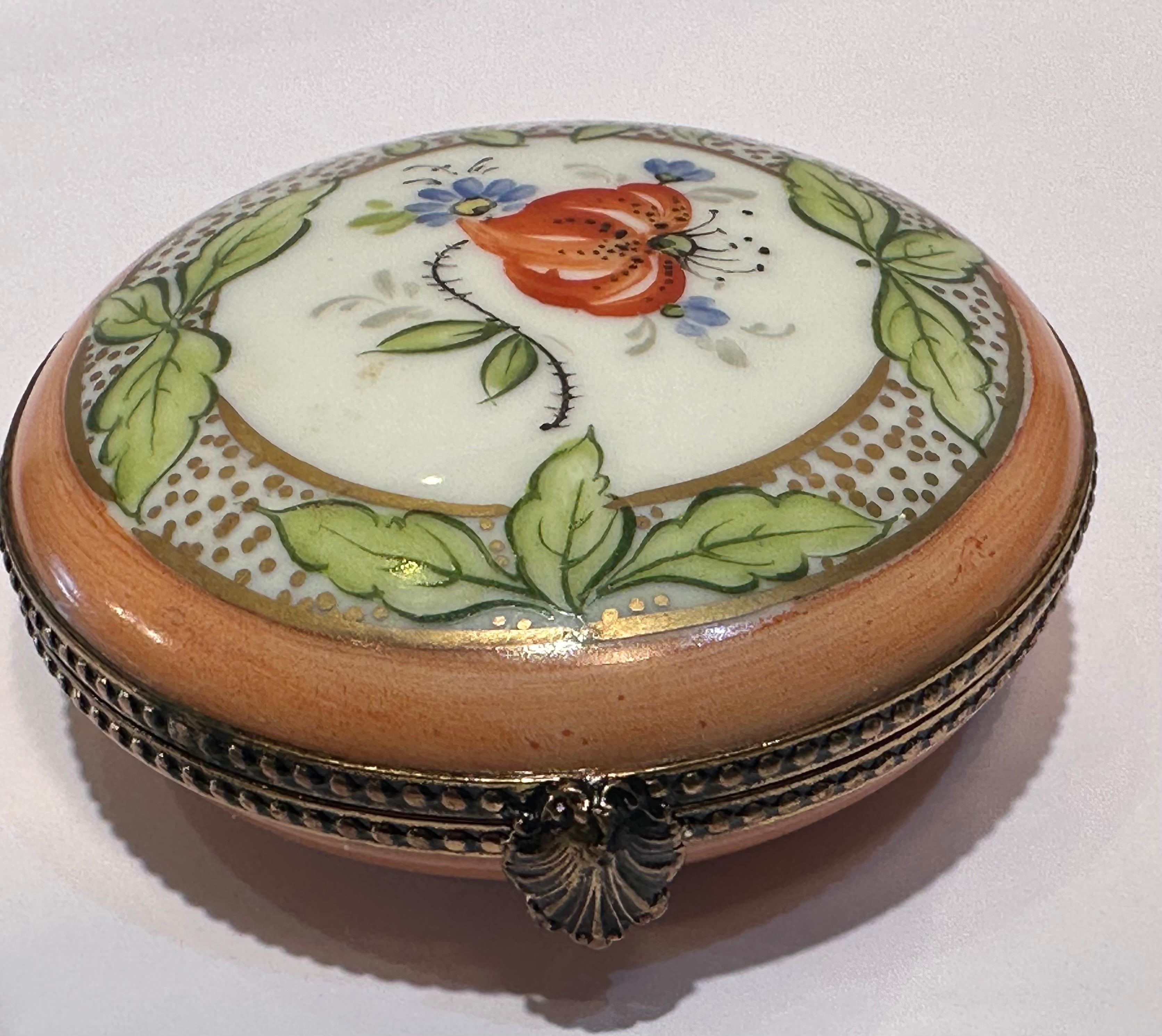 Beautiful Limoges porcelain circular shaped trinket box is handmade with a peach and white background and is skillfully hand painted with a floral and leaf motif and richly accented with 24-karat gold dots and trim. Box features antiqued metal