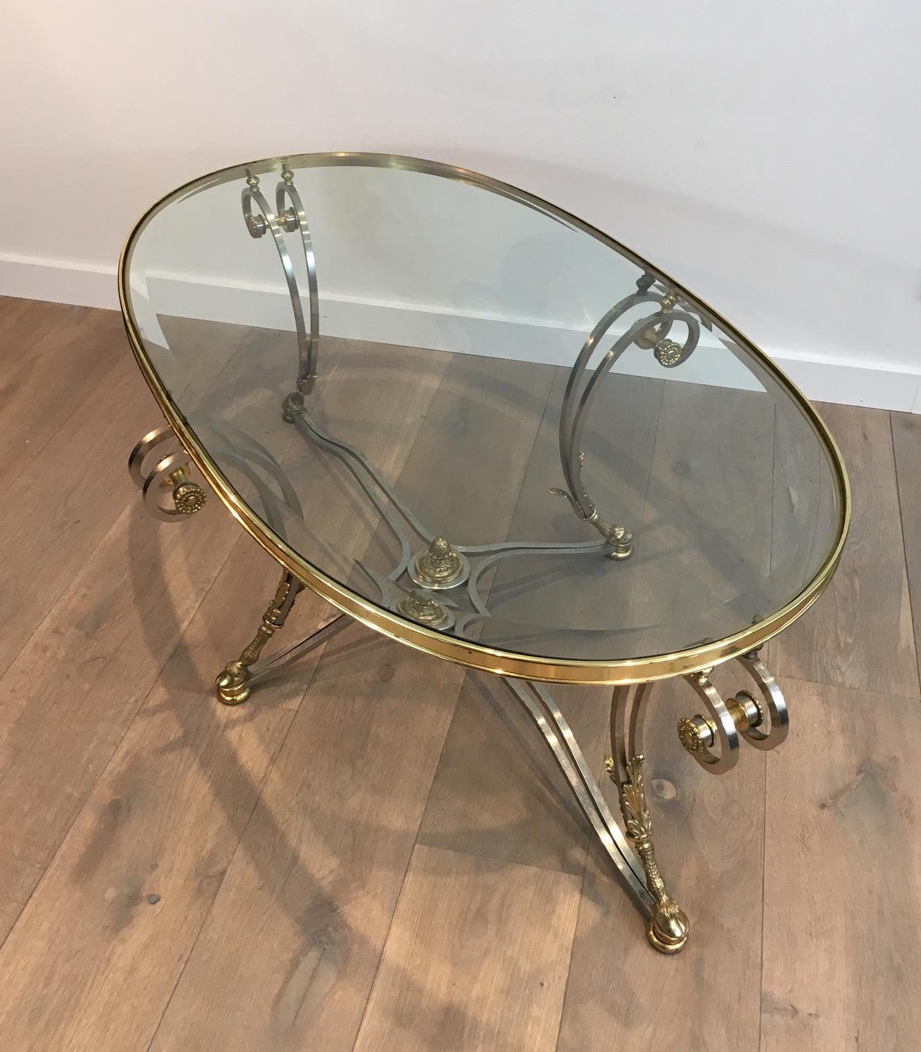 This beautiful neoclassical oval coffee table is made of brushed steel and brass with a nice oval beveled glass top. It has nice ornaments, a very elegant stretcher with a finial on its center and four brass down feet. This is a delicate work in the