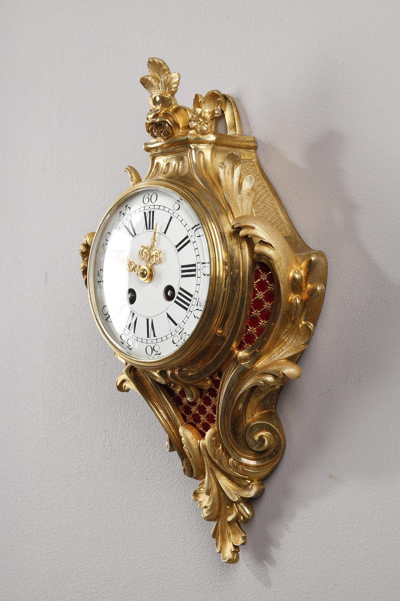 Small gilded bronze cartel. Its rocaille decoration of animated foliage is of Louis XV style. It is adorned with an openwork pattern that uncovers a red velvet fabric. The dial is enamelled with Roman numerals for the hours and Arabic numerals for