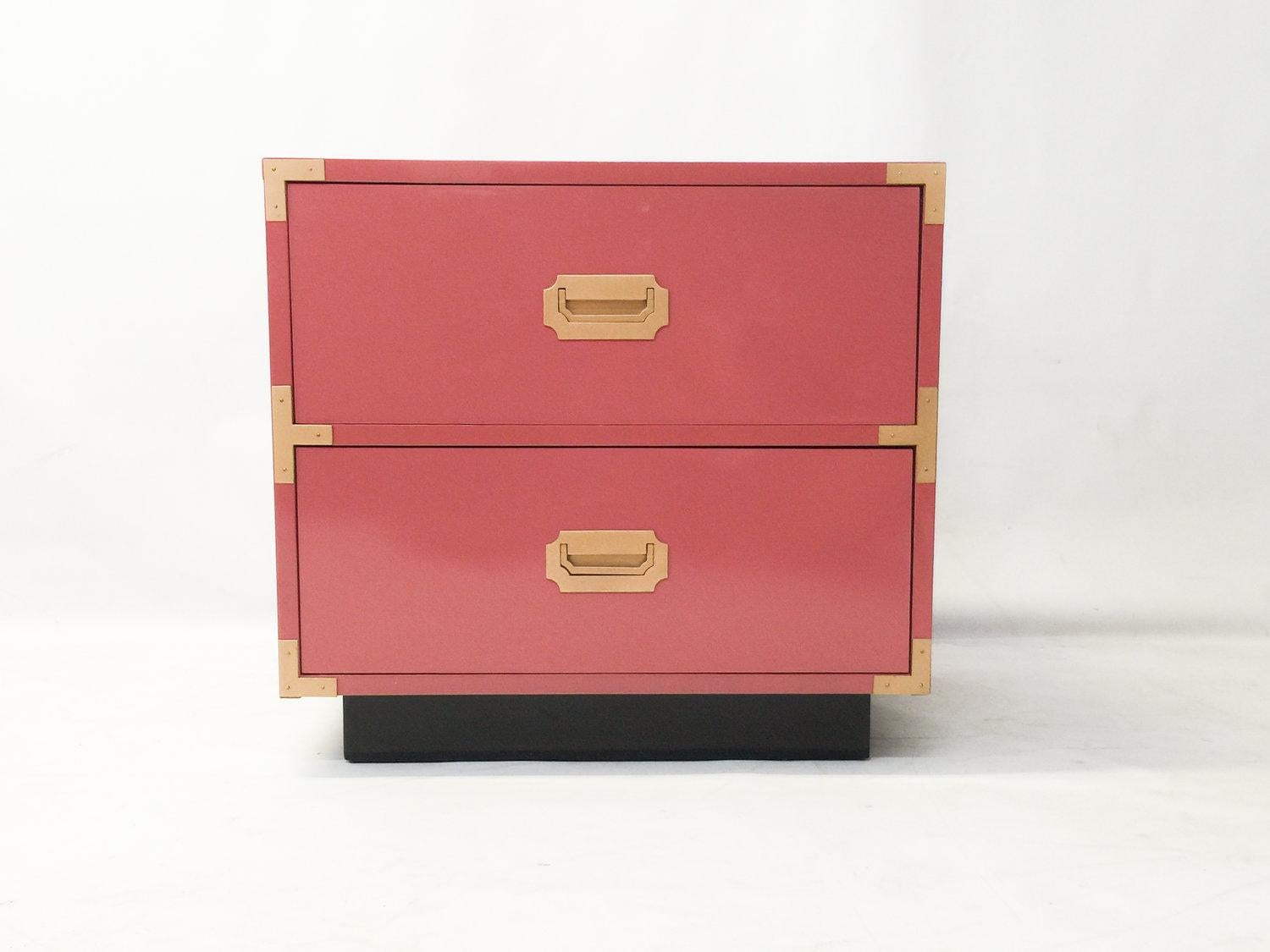 A beautiful pair of Mid-Century Modern Campaign style nightstands. These have been professionally lacquered in pink with a black base. Solid wood construction these feature plenty of storage, two drawers with recessed polished brass pulls resting on
