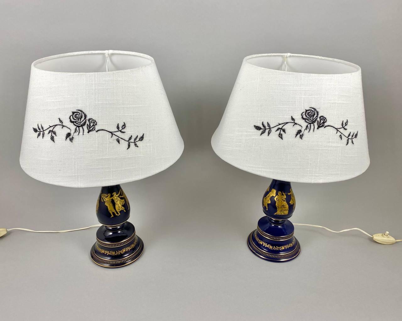 A lovely pair of porcelain cobalt blue table lamps with Gold Greek Mythology paintings and brass fittings. 

 The lamps will find its place in the living room and in your bedroom.

 The lampshades are made of white fabric with floral