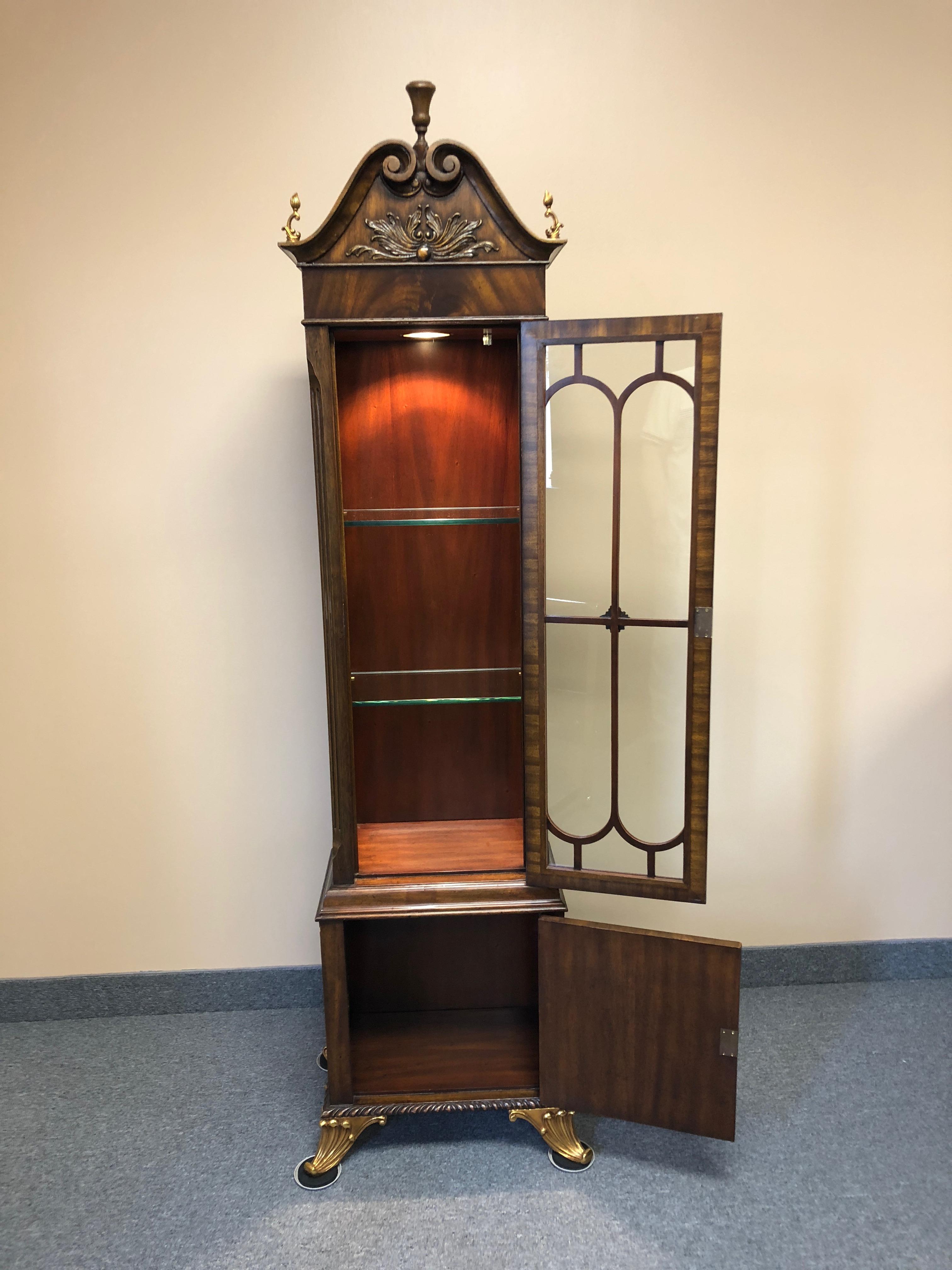 A beautiful burl mahogany display cabinet having gilded brass embellishments, a paned glass door that opens to adjustable glass shelves and interior lighting, a single inlaid panel door beneath with storage inside, and gorgeous decorative feet.
