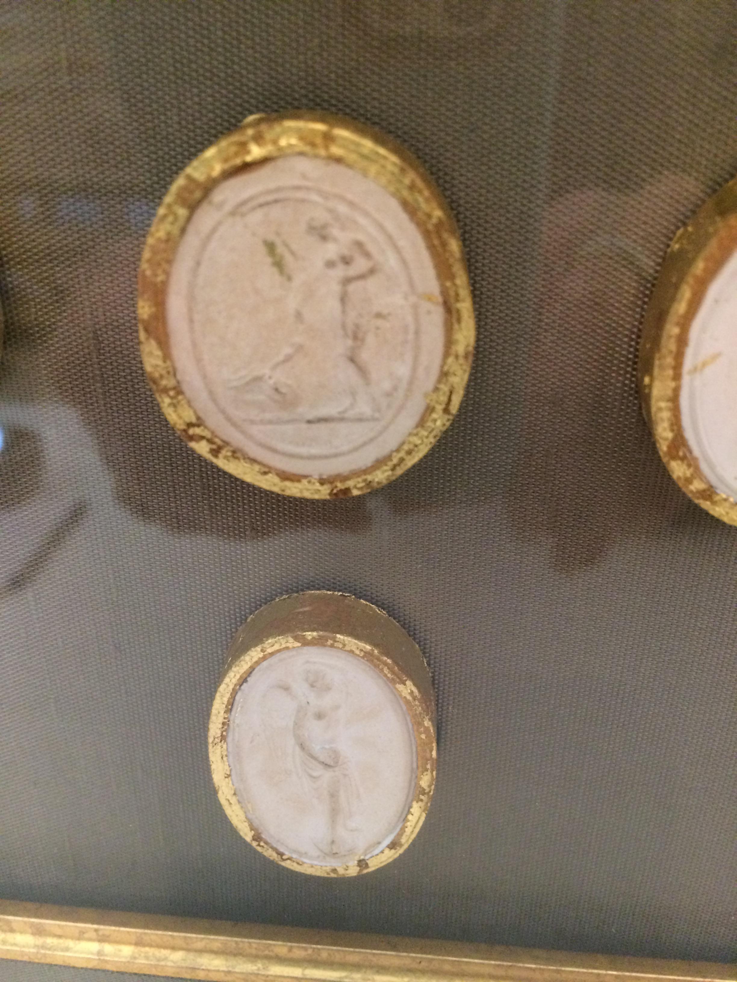 A set of five cream colored coin shaped intaglios encased in gold leaf and beautifully framed against a taupe silk background, double matted with gold leaf and a gilded frame.