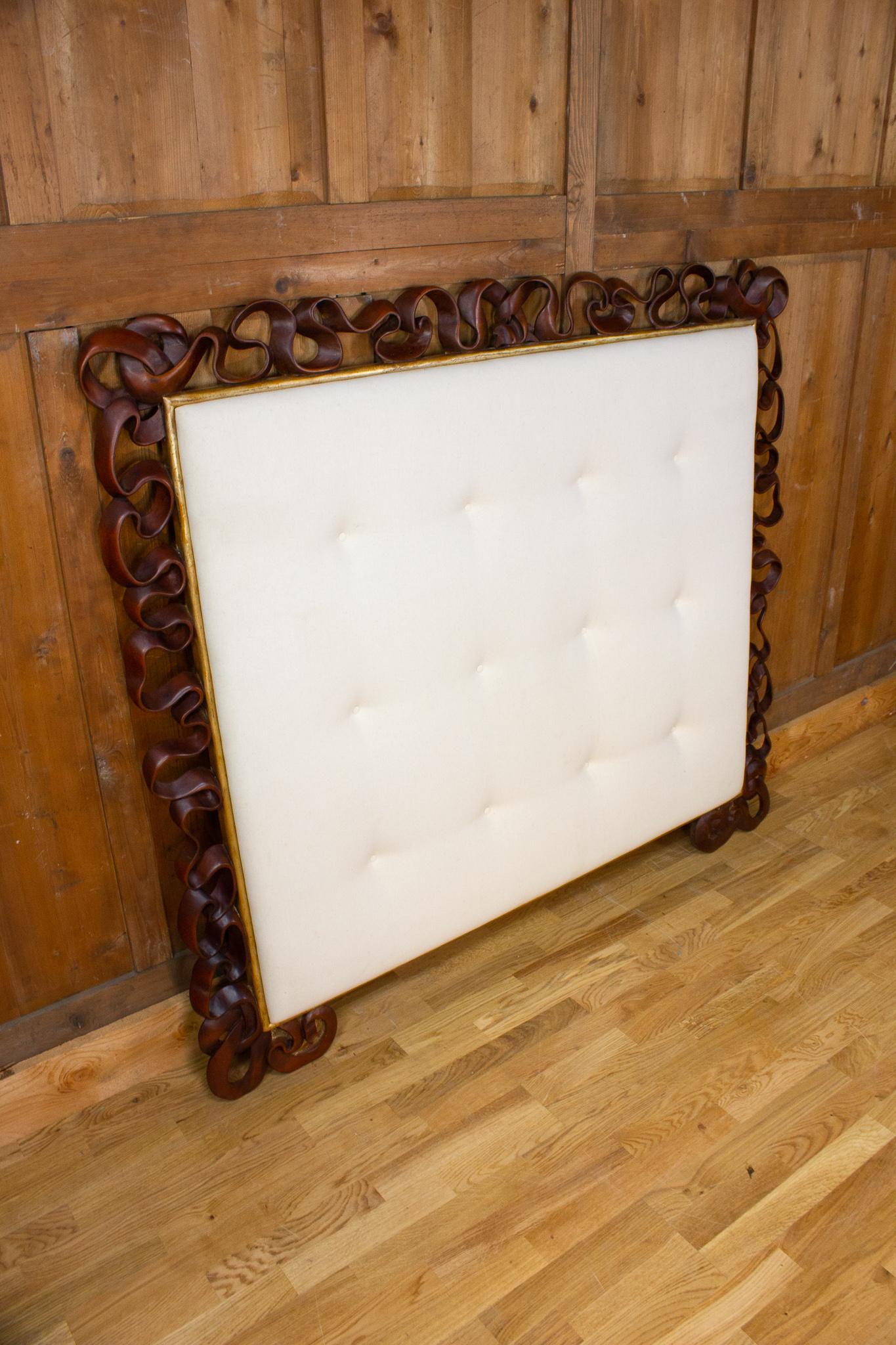 Beautiful and very decorative headboard in carved wood and fabric. The exotic wood structure, most likely mahogany, is carved with a ribbon motif. The white padded fabric is framed by a gilt wood molding. 
Probably Italy 20th century.