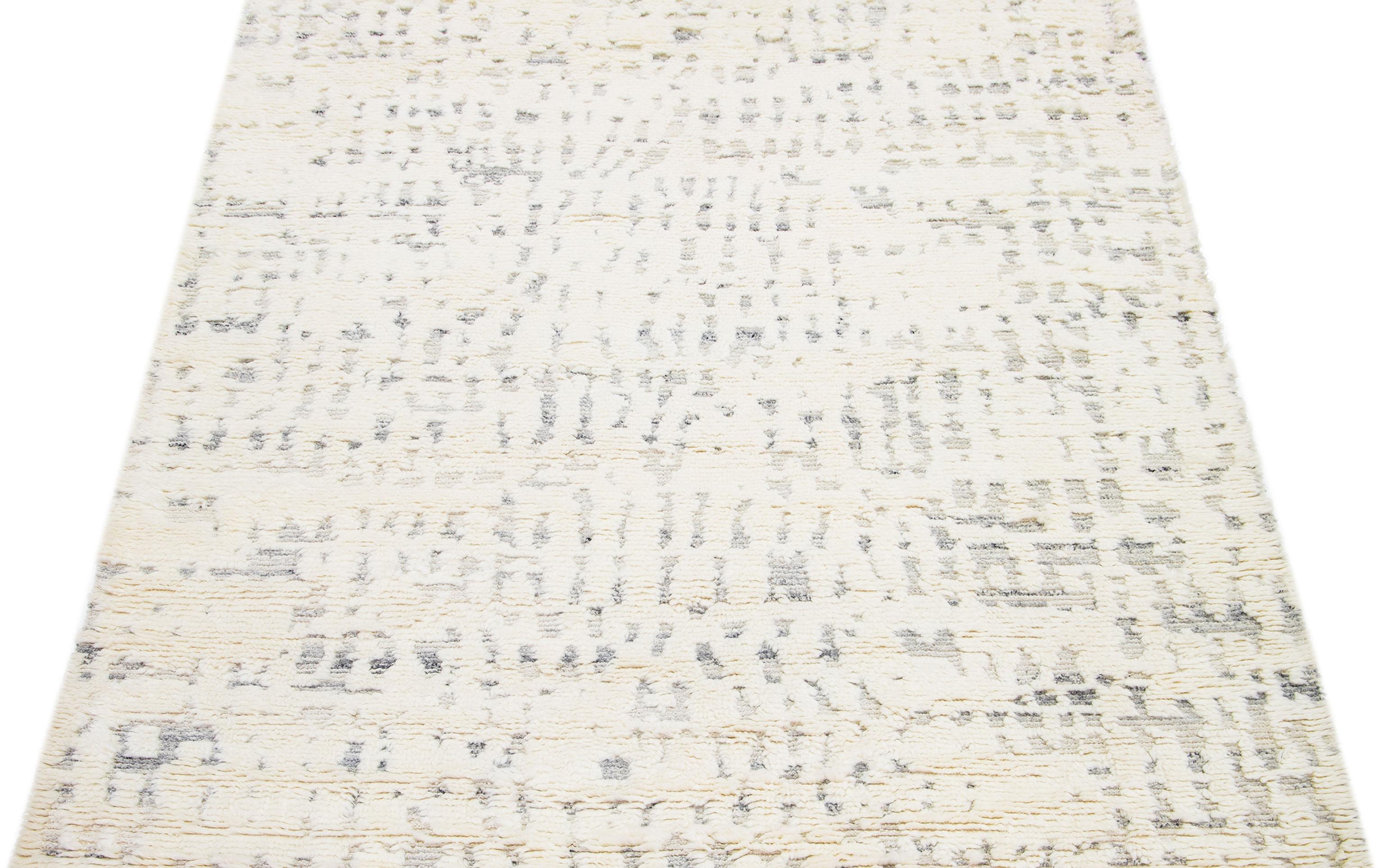 Esta alfombra de lana finamente elaborada a mano muestra un elegante motivo marroquí meticulosamente tejido en tonos grises, que proporciona un exquisito contraste con su encantador lienzo marfil. La alfombra presenta una estética abstracta que