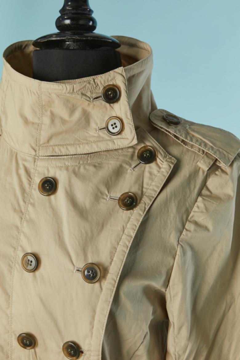 Beige cotton trench coat with asymmetrical button placket. Pocket on both side. Belt in the same fabric with buckle and belt-loop. Flap on the top of the back with buttoin and buttonhole closure. One button and buttonhole on the middle bottom back