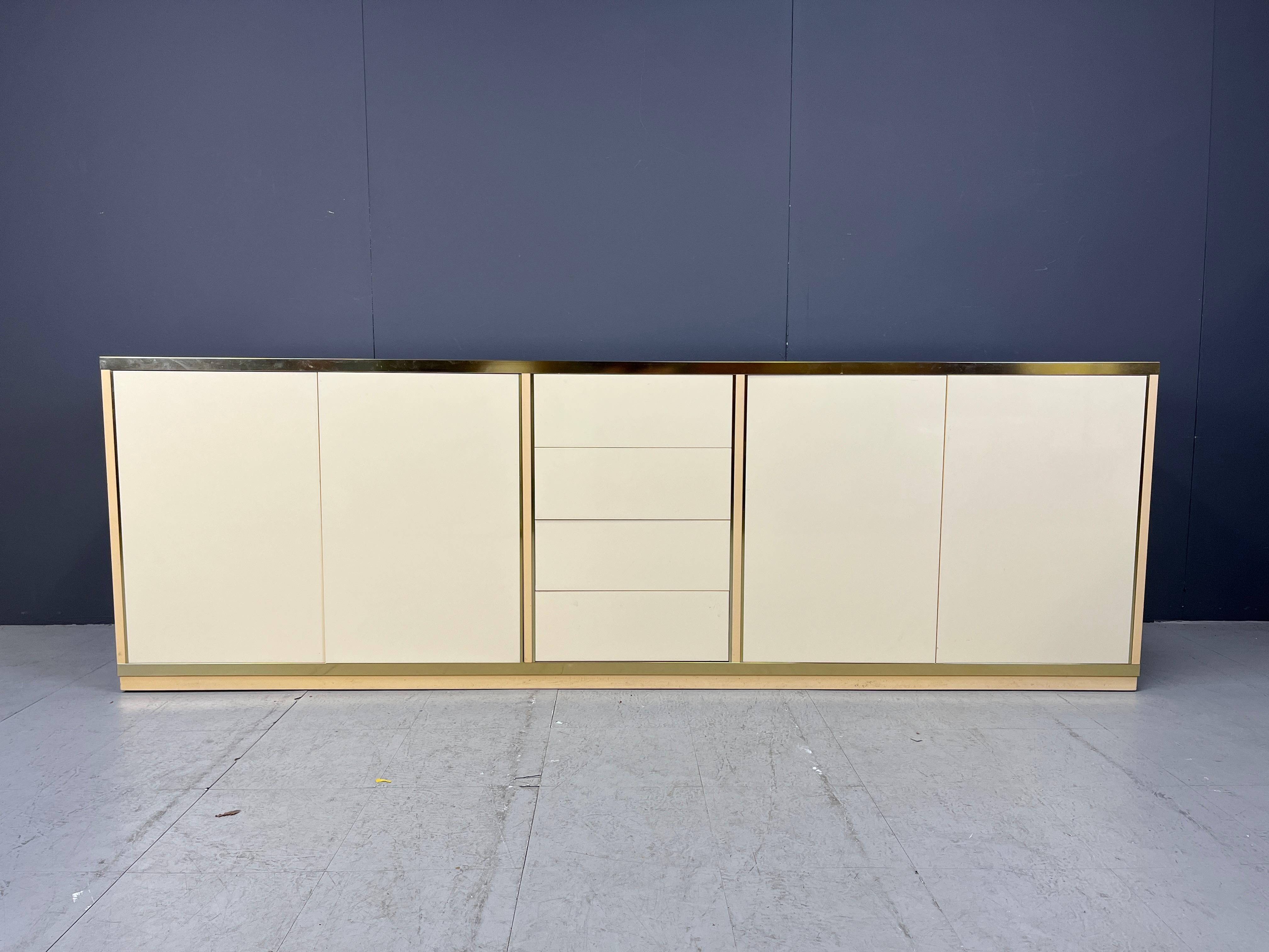 La crédence est fabriquée en bois laqué beige et en bandes de laiton.

Il comporte quatre tiroirs et quatre portes qui révèlent un grand espace de rangement.

Dans l'ensemble, cette belle pièce est un véritable accroche-regard et un bel exemple de