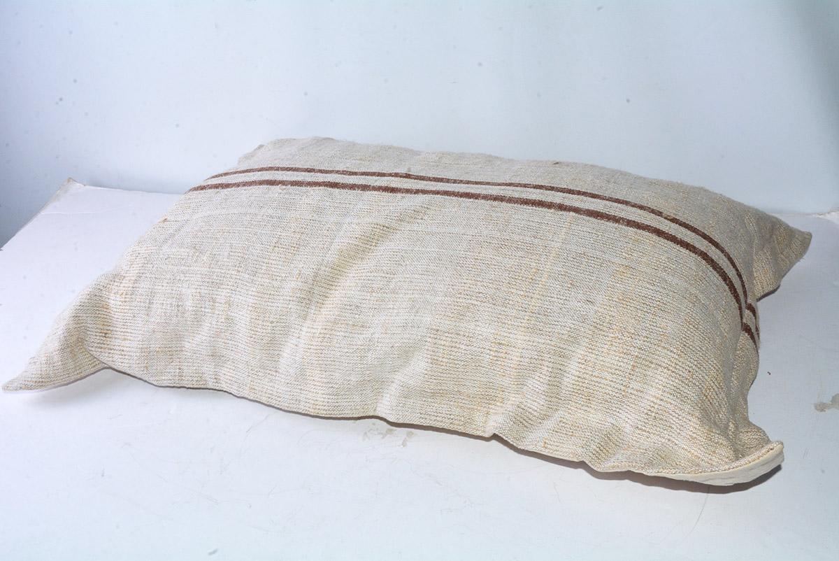 The beige grain sack linen pillow cover with a rustic texture and brown woven double stripes encases an inner cotton pillow. Two shell buttons on the back secures the cover. The back is made of a finer woven beige linen. An encircling phlange