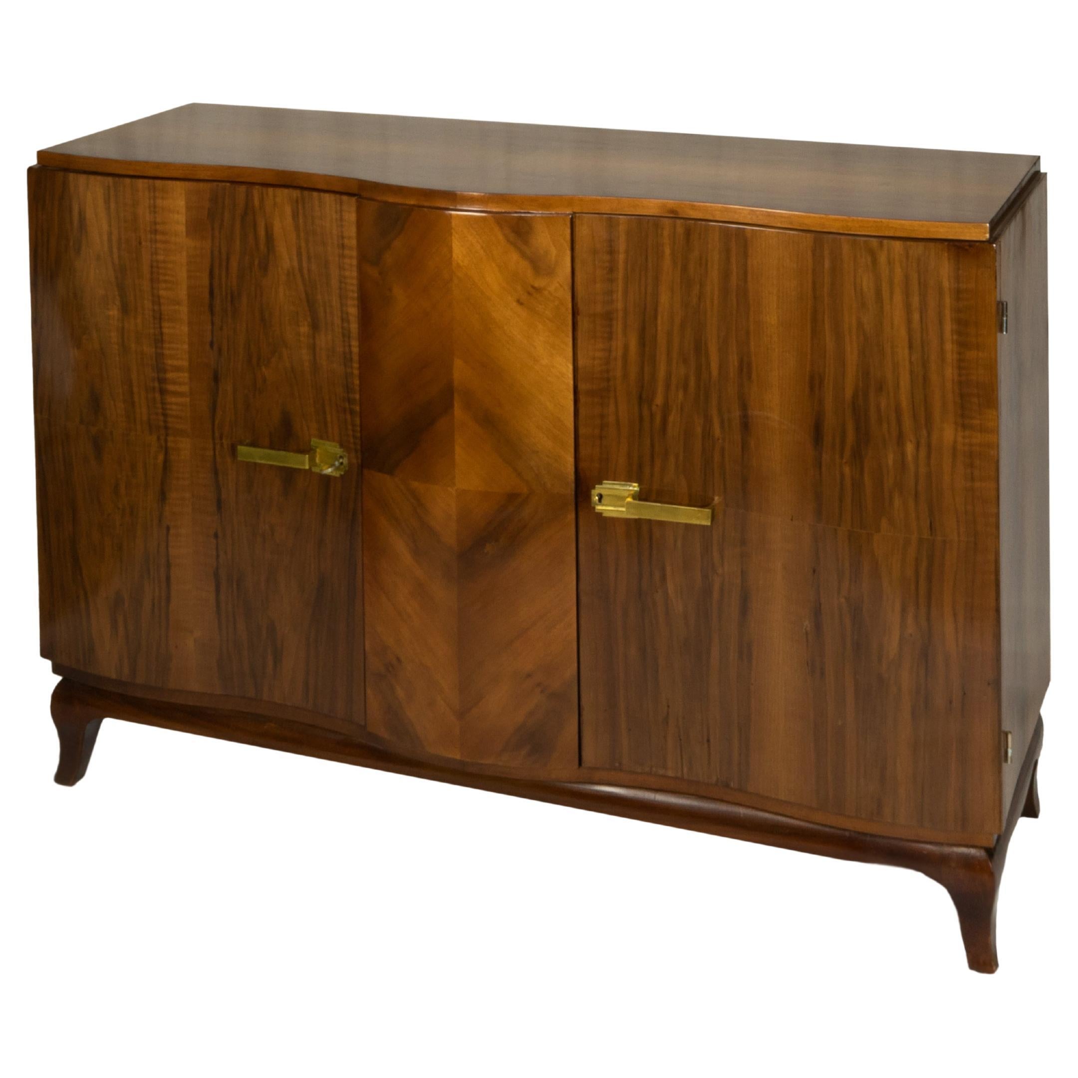 This Belgian Art Deco sideboard is crafted from walnut and stands on four flared legs. 
The sideboard features two doors with golden metal hardware and offers a stylish storage solution for apartments or modern spaces. 

It’s a fine example of 20th