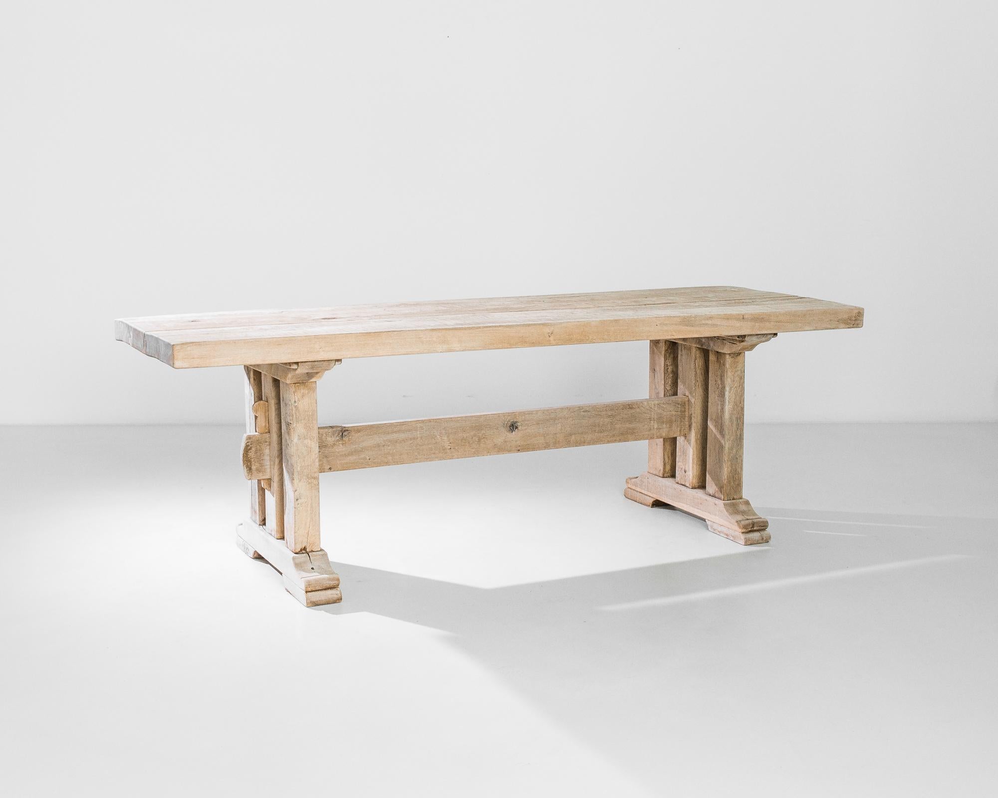 An oak dining table from 1960s Belgium with a handsome farmhouse silhouette. A long tabletop of wooden girders sits atop trestle legs, joined with a strut. The contours of the feet and pegs lend a shapely inflection to the sturdy form. The oak has