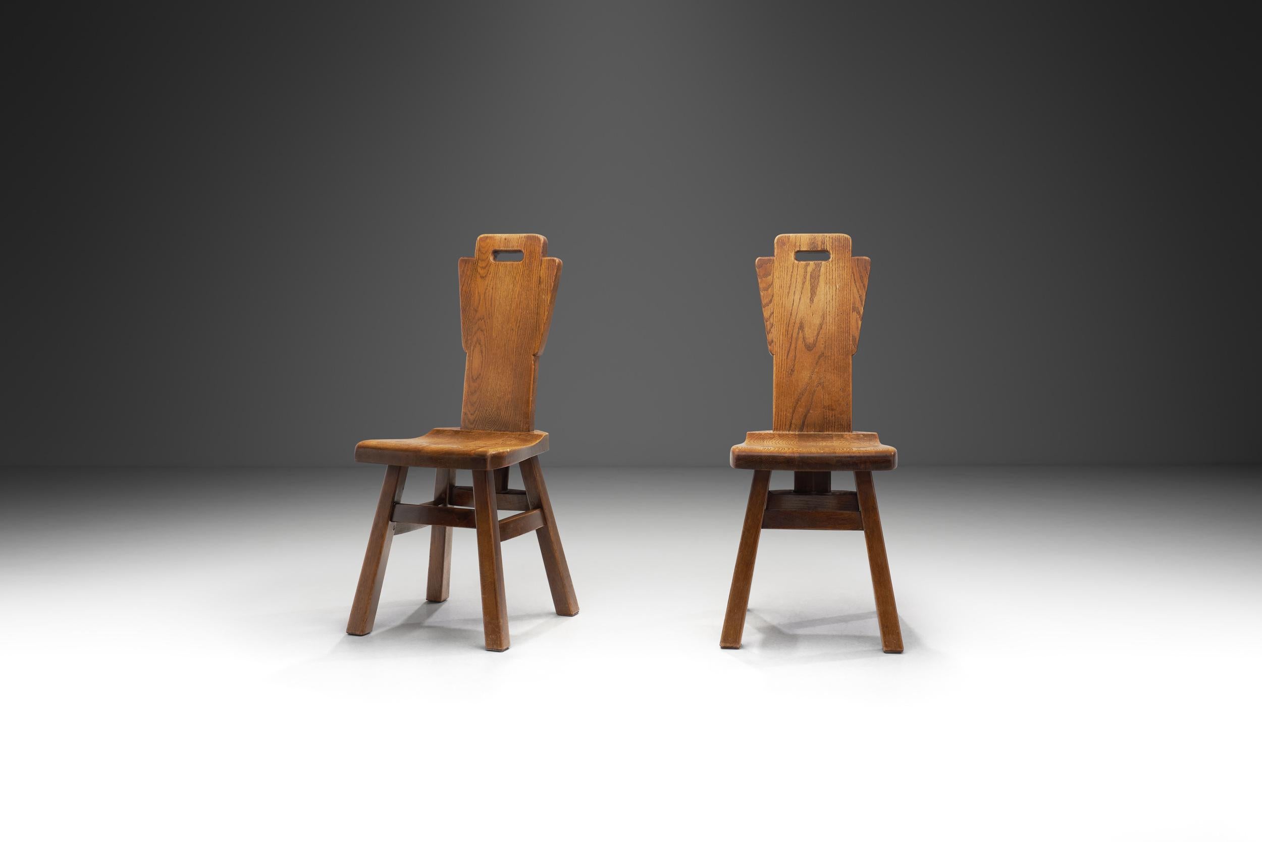 In the tumultuous design landscape of mid-20th century Belgium, this pair of chairs emerged from the hands of skilled craftsmen during the 1970s. Crafted from the dense and unyielding embrace of heavy oak, these brutalist pieces stand as Stoic