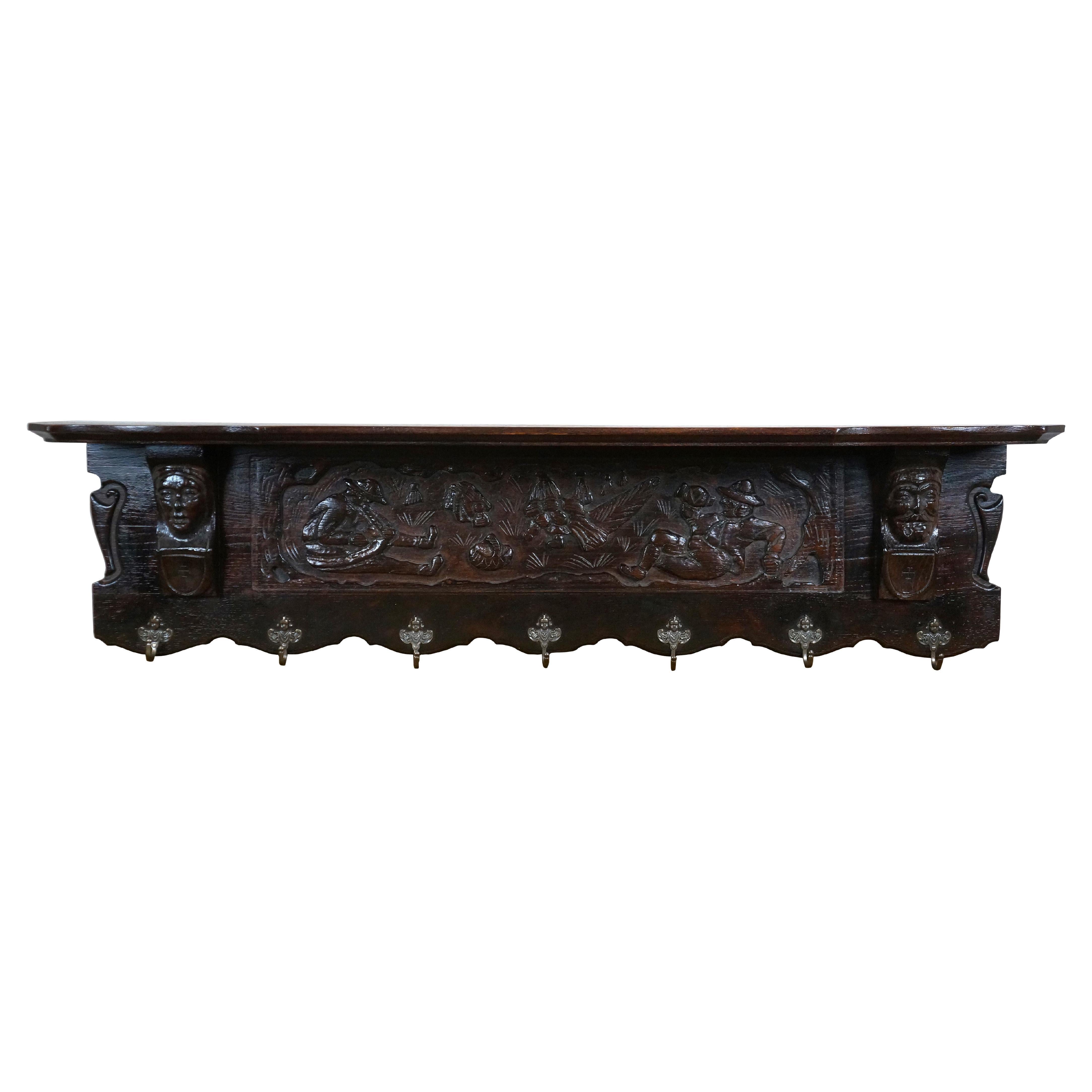 Albero da parete in legno belga intagliato con sette ganci a forma di cherubino / angelo / putti per cappotti / cappelli. La scena intagliata raffigura una coppia di cacciatori seduti per riposare con un corno e un boccale. Un ripiano superiore con