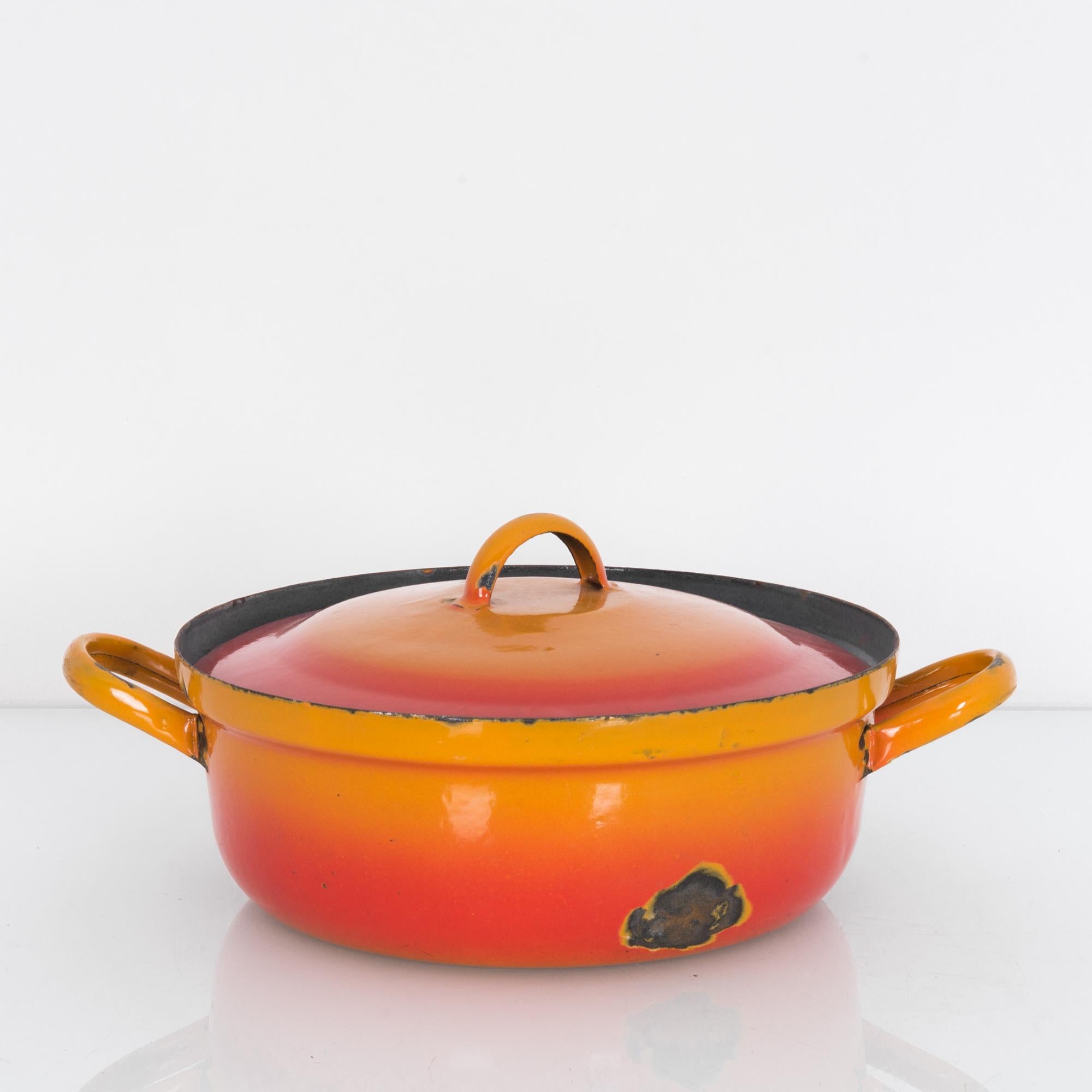 A metal cooking pot from Belgium. An ample casserole-style pot with looping handles and a low, bell-shaped lid. The enamel finish is gradated in bright sunset tones — from deep orange to light peach. Worn patches on the base and rim lend a warm,