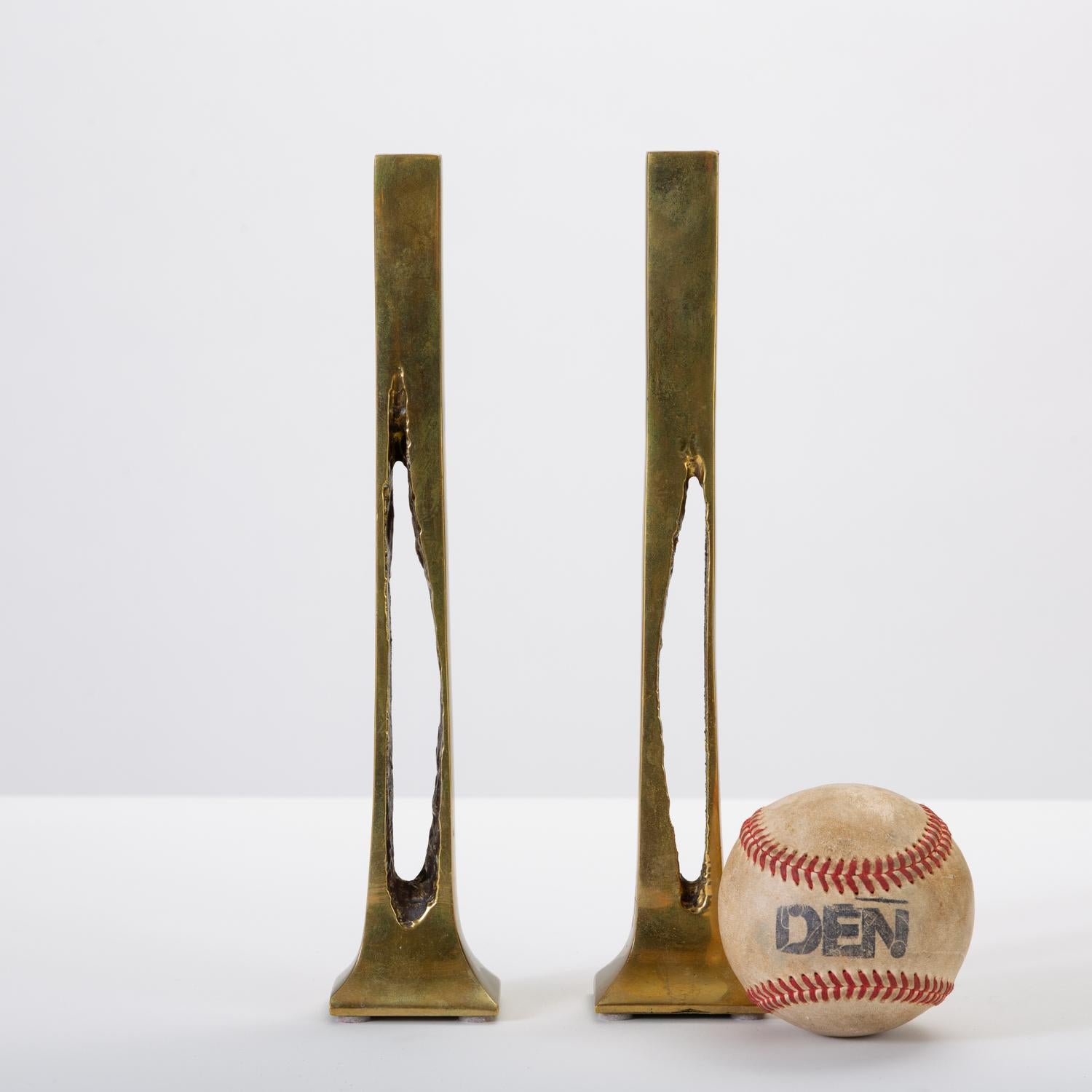 A pair of cast brass candlesticks from 1970s, Belgium. The slender silhouette has a sculptural body with irregular negative space at the centre, widening at the foot to a rectangular base. The outer brass is lacquered with a glossy finish; the