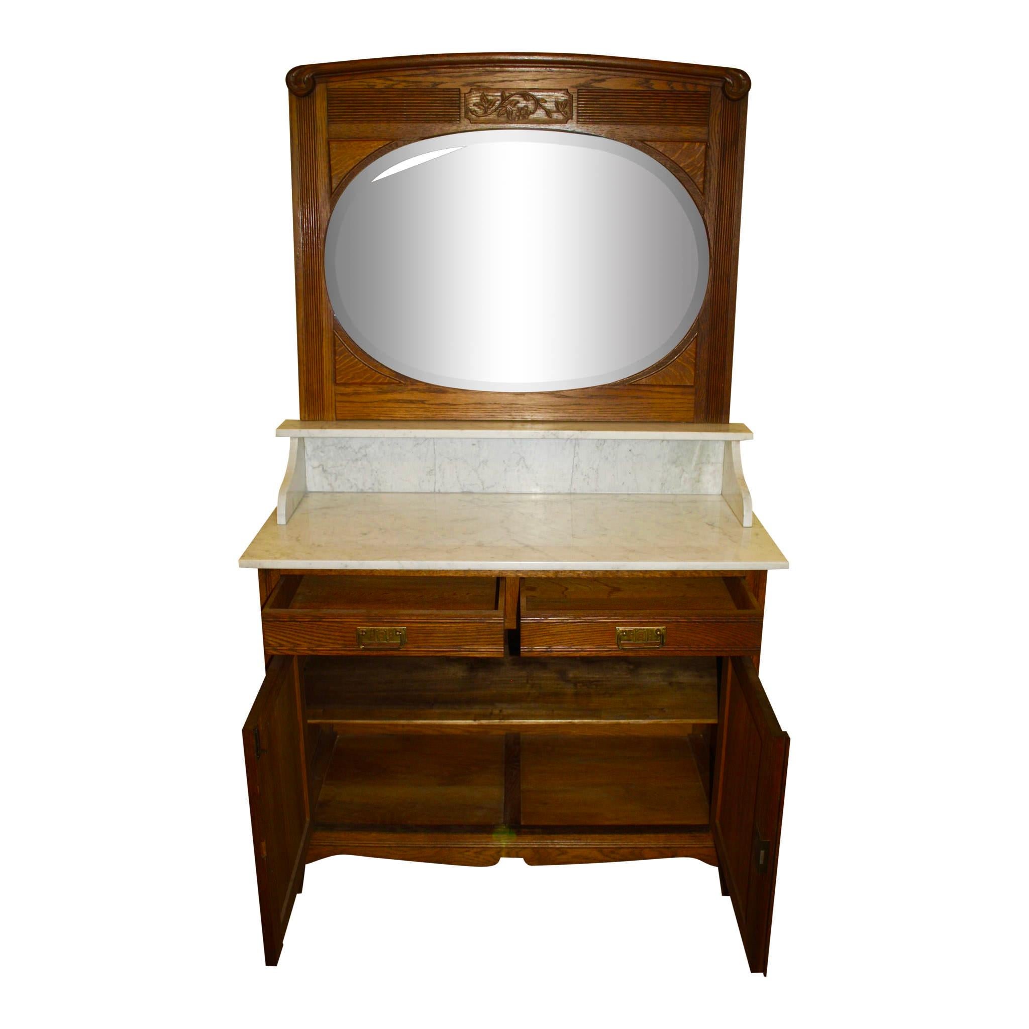 A beveled, oval mirror framed in oak with fluting and a floral carving sits above the gray veined marble countertop of this Belgian vanity dresser. Convenient storage is provided in the two drawers and floral carved double doors, which open to a