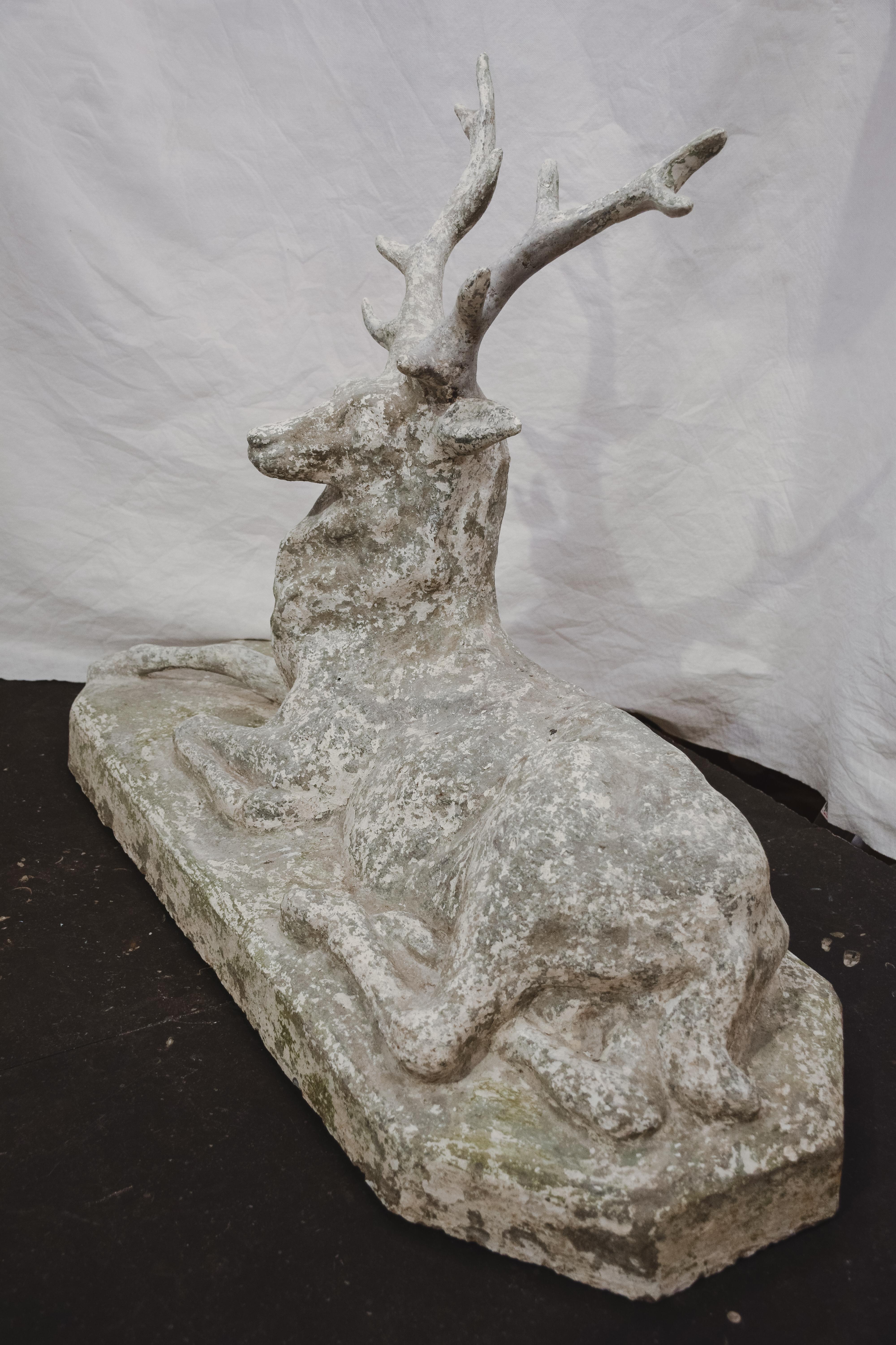 A reconstituted stone reclining stag sculpture from the early 20th century, with nicely weathered patina. The sculpture depicts a stag laying on a conforming base. Boasting a nicely weathered appearance revealing its age and life outdoors, this