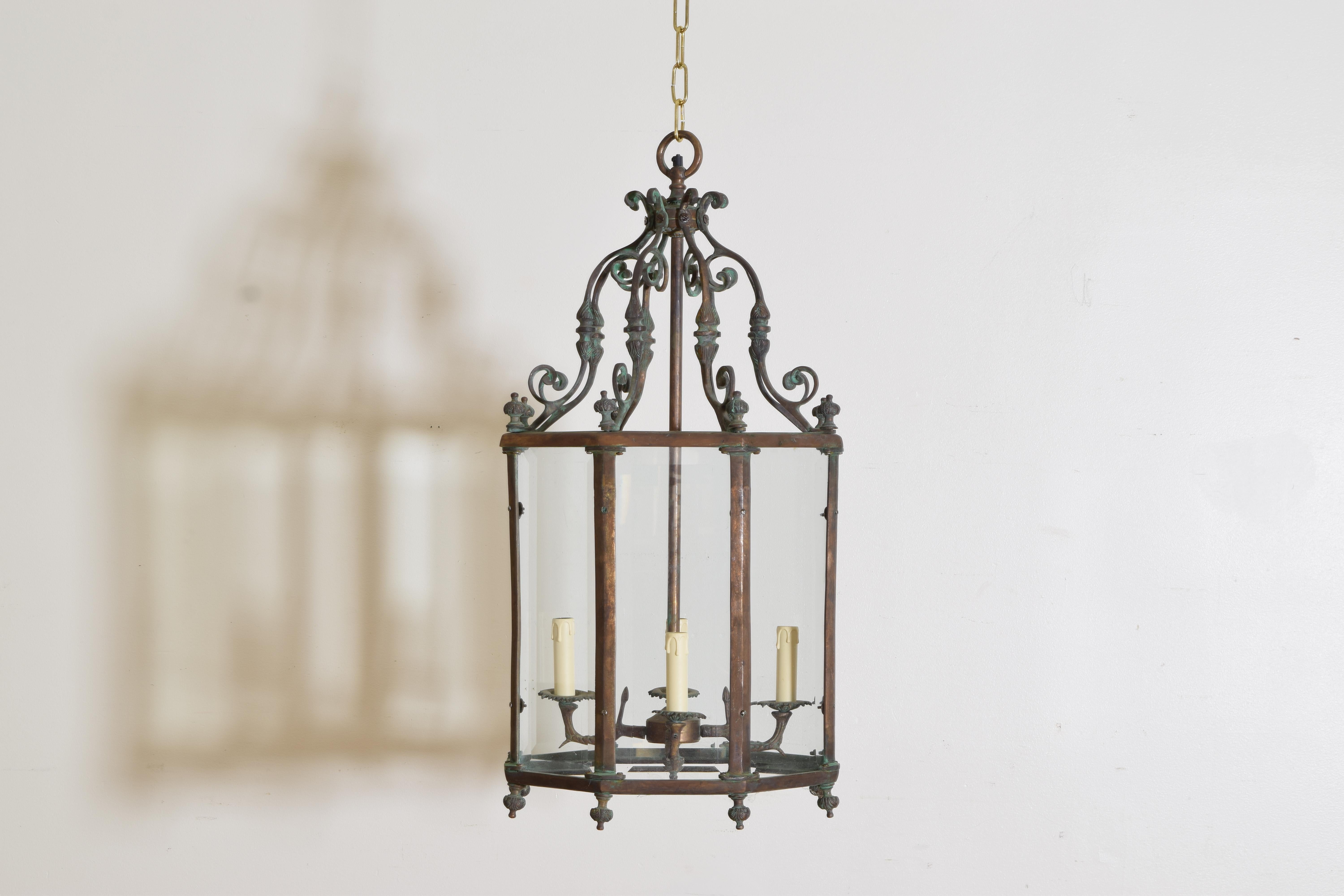 Hanging from a ring above a framework of brass scrolls, the body of this lantern is constructed of brass and copper and it houses individual panes of beveled glass, retaining its original cluster of four lights, with brass “feet” at intersections of