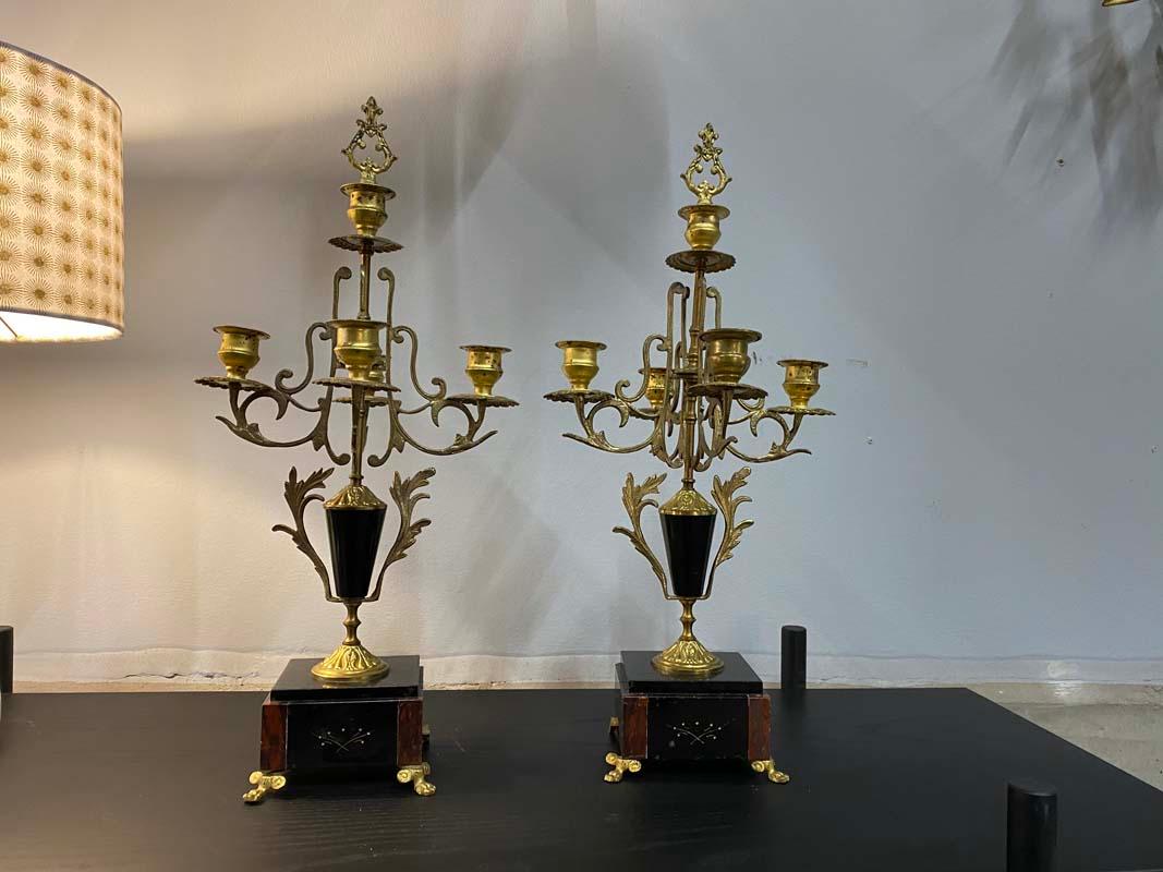 These two candlesticks are from the beautiful years of the turn of the 19th century into the 20th century in France, the so-called Belle Epoch. 

The golden intricately designed upper part of the candlesticks is made of brass, the lower plinth is